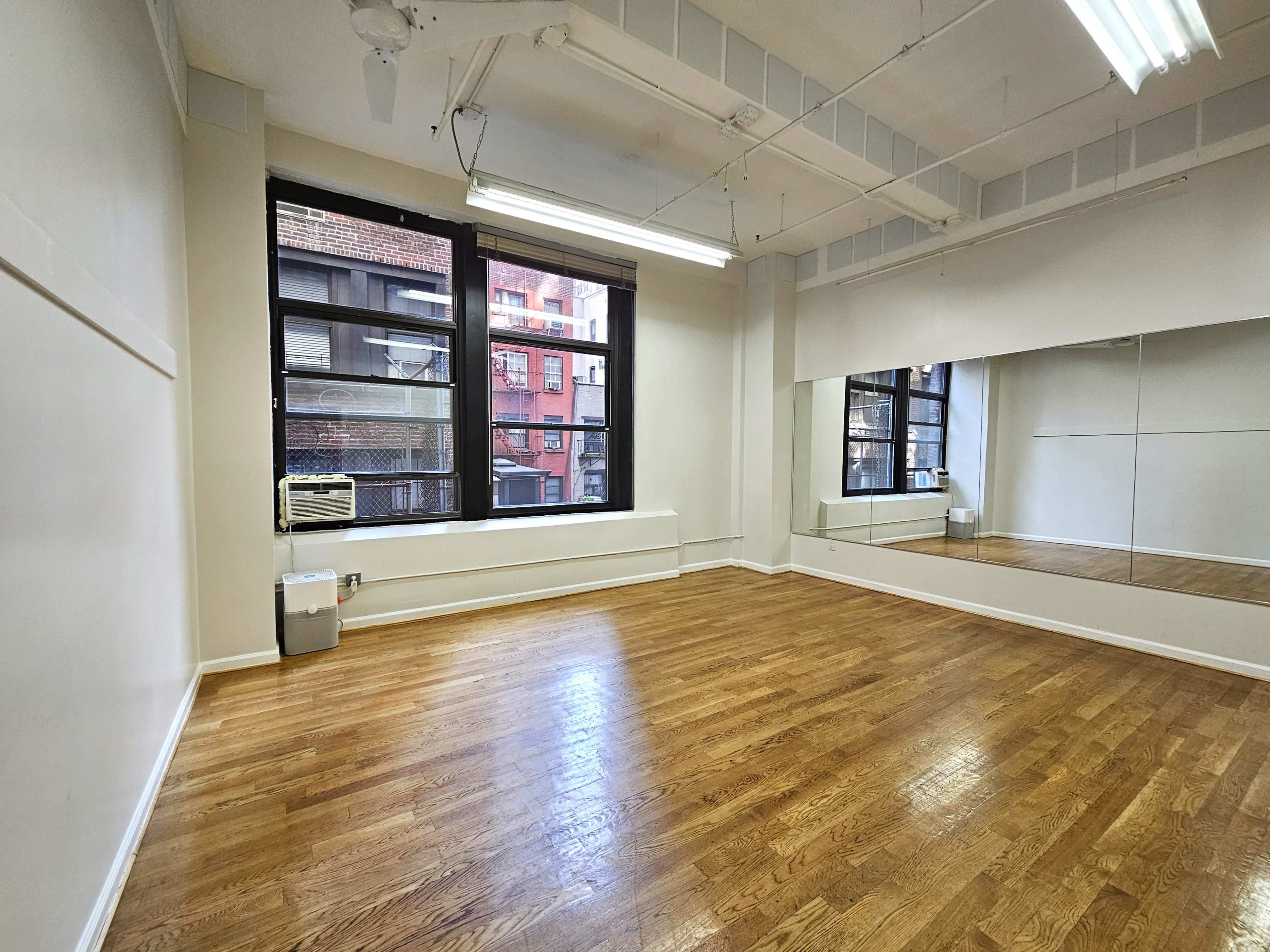 An empty room with wooden flooring, large windows on one side, and a mirror along one wall.