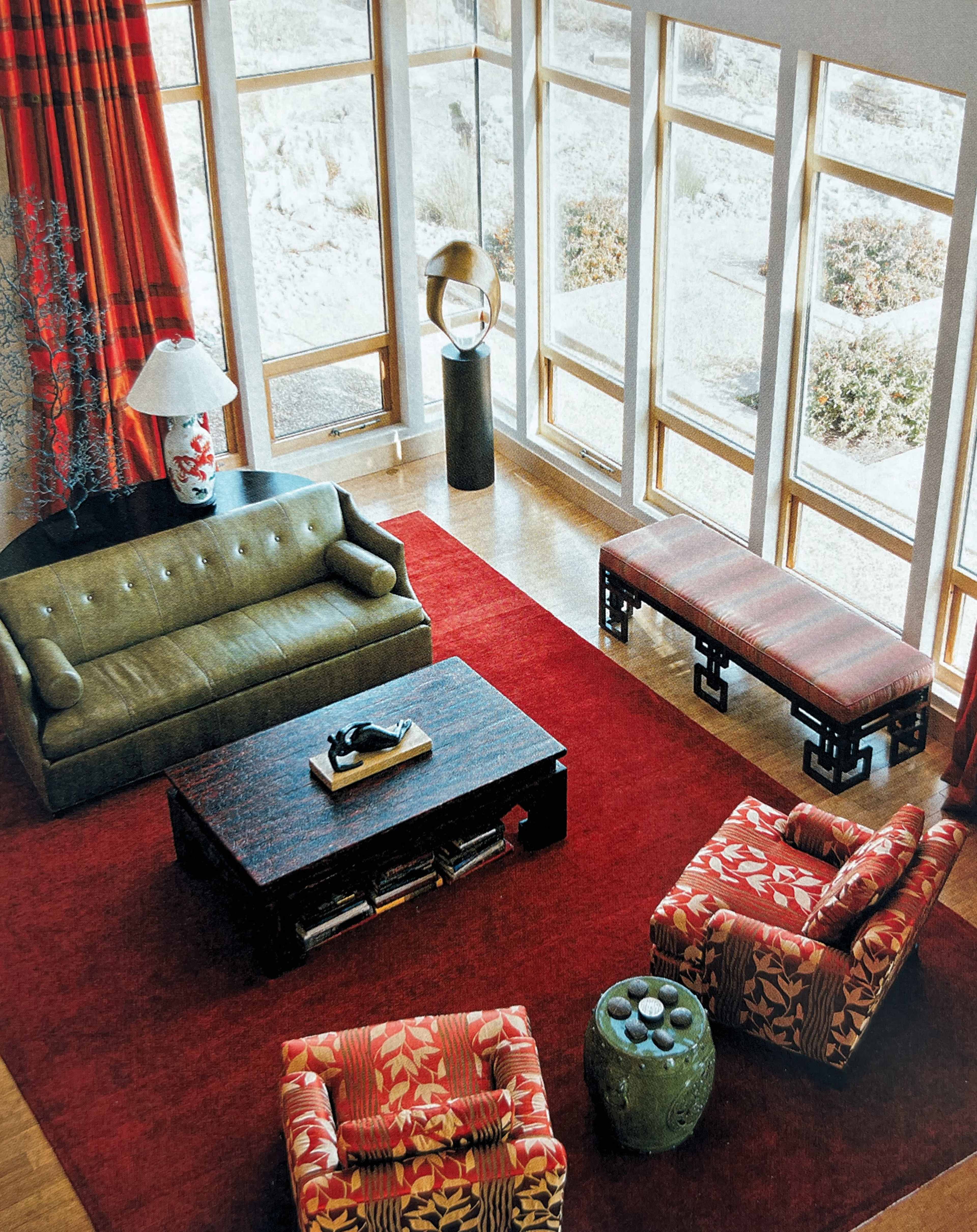 The image depicts a modern living room with large windows, a green sofa, patterned armchairs, a coffee table, and a red area rug.