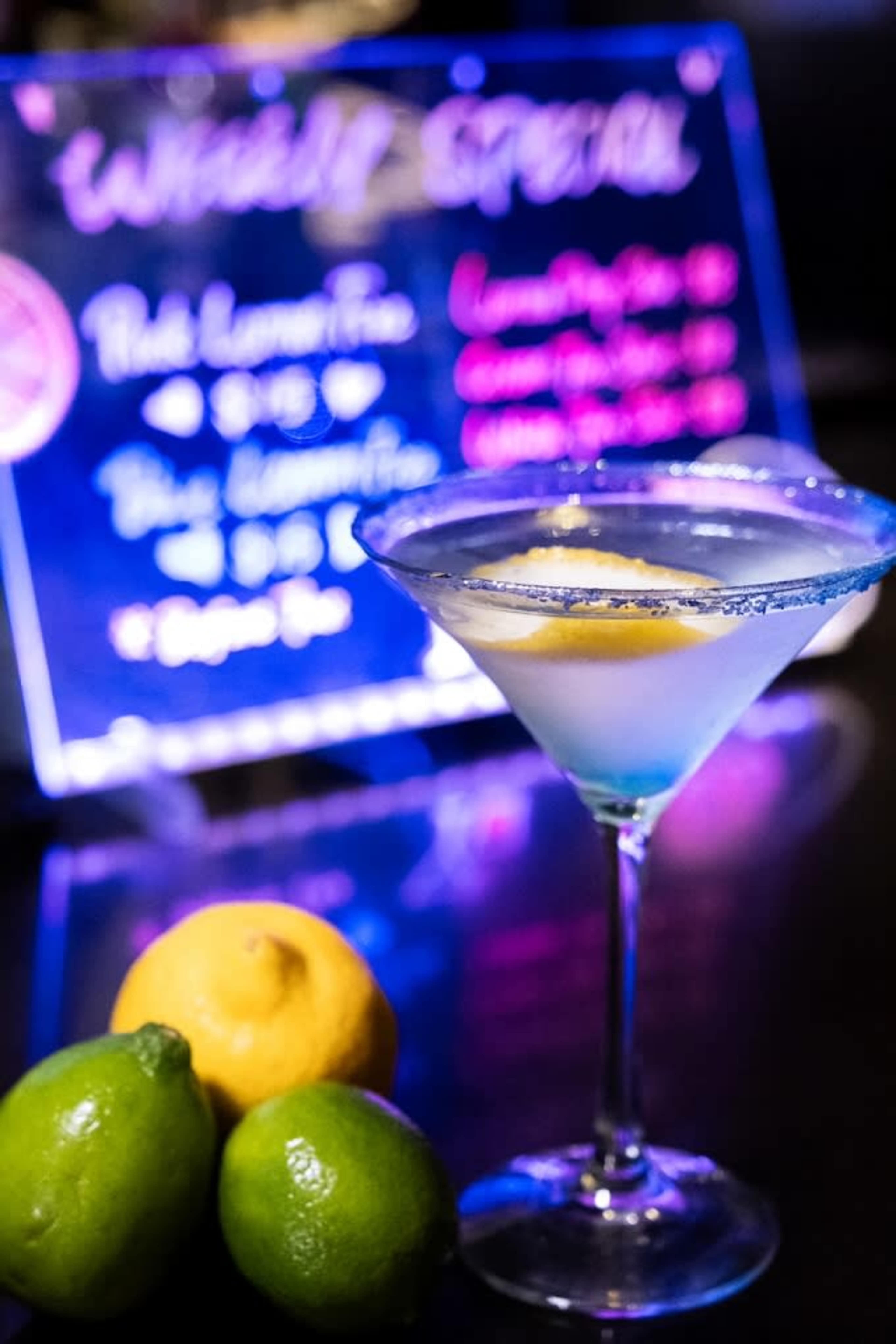 A cocktail glass with a lemon twist sits in front of a menu board featuring daily specials, along with a lemon and two limes.