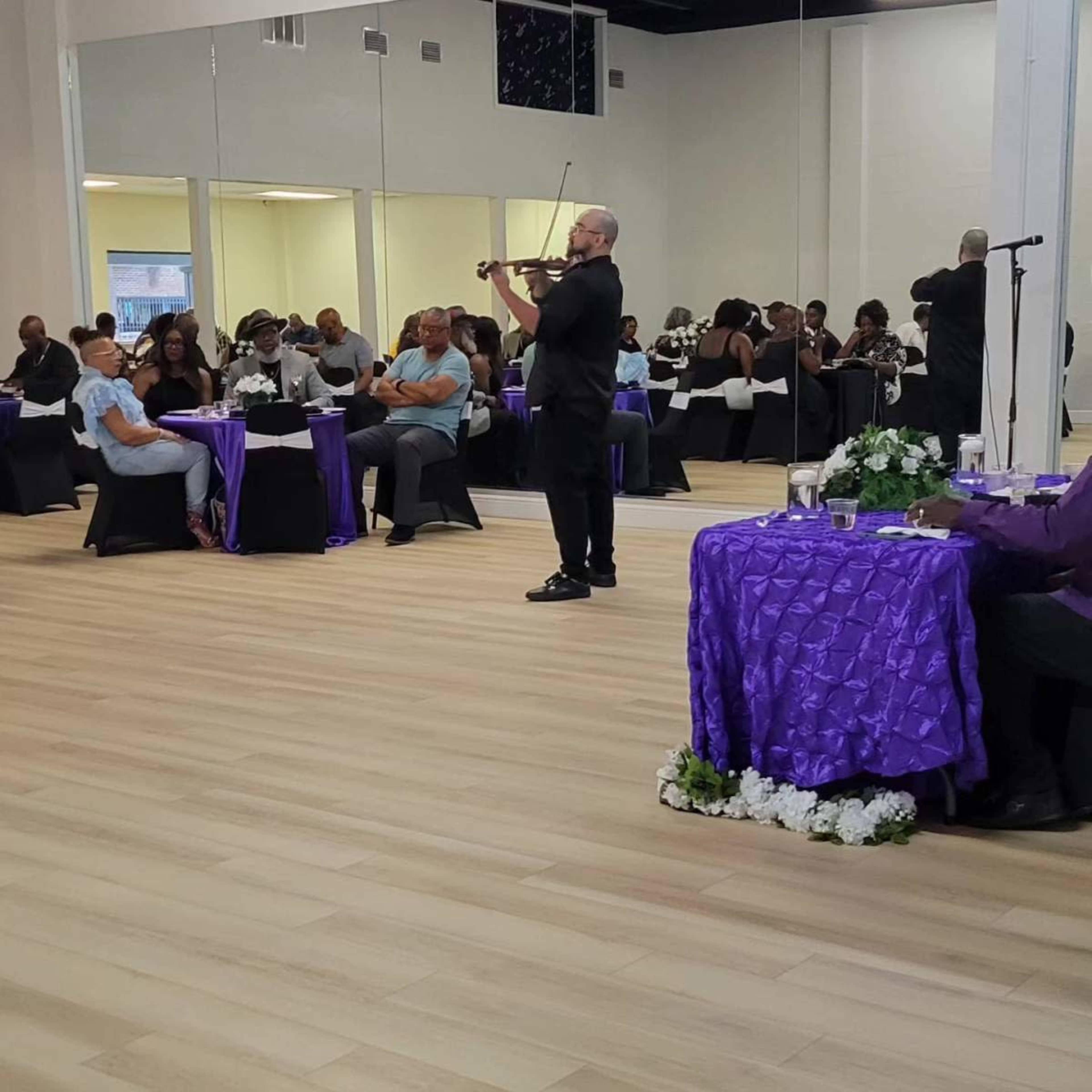 A man plays the violin in a banquet hall filled with seated guests at elegantly set tables.
