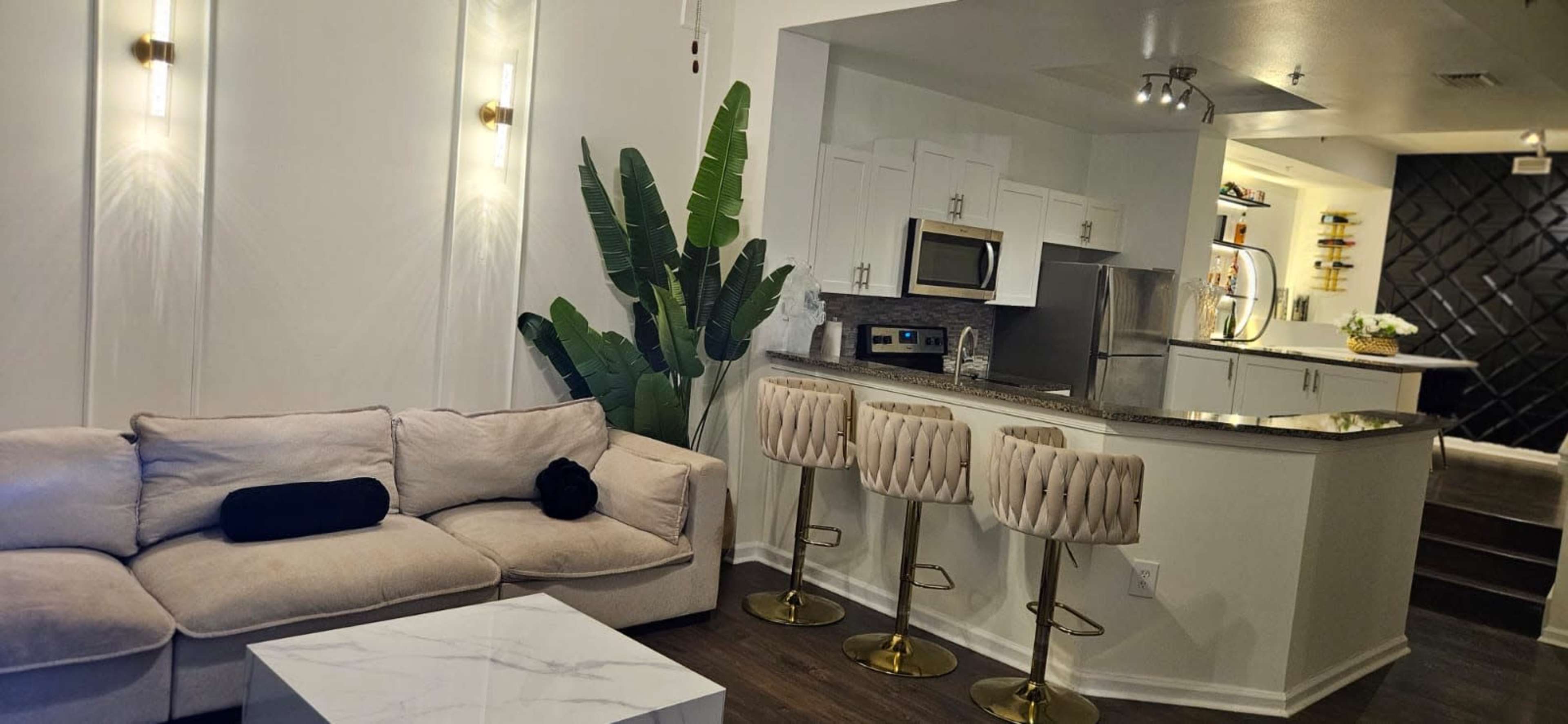 The image displays a modern living area with a beige couch, a white marble coffee table, and a kitchen in the background featuring stainless steel appliances and gold-accented bar stools.