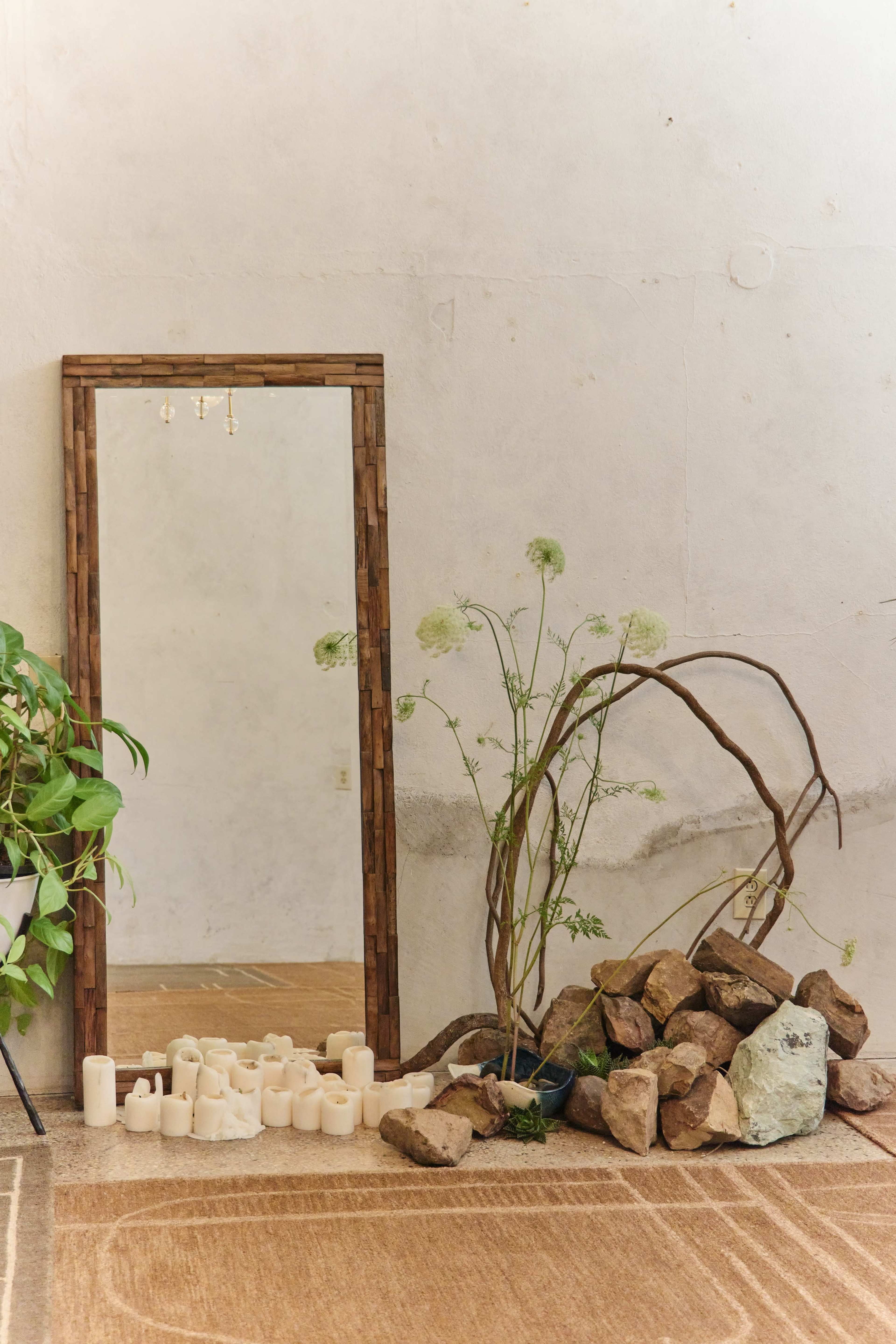 A large wooden-framed mirror leans against a textured wall beside a collection of rocks, dried branches, and several candles arranged on the floor.