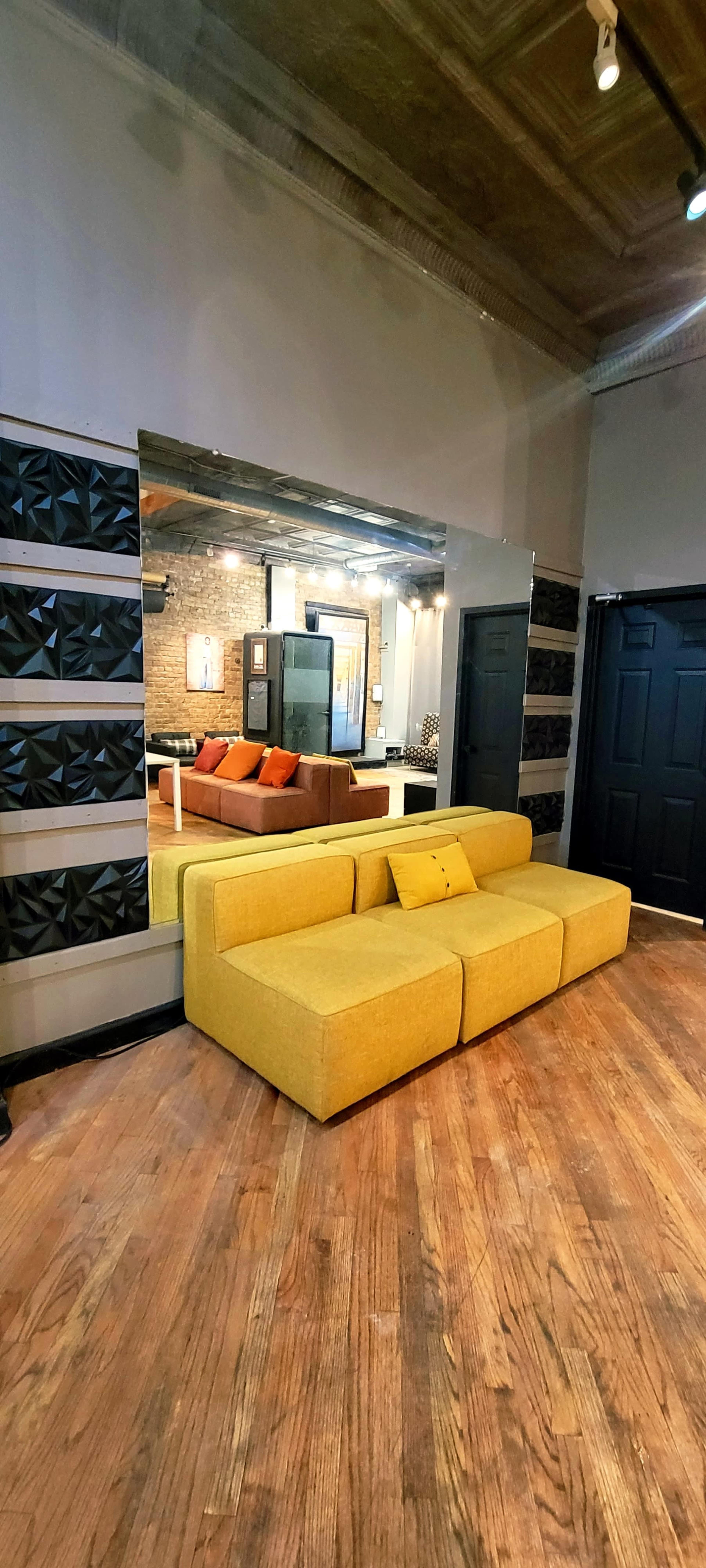 The image shows a modern interior featuring a yellow modular sofa in front of a large mirror, with a glimpse of an orange and brown seating area visible in the reflection.