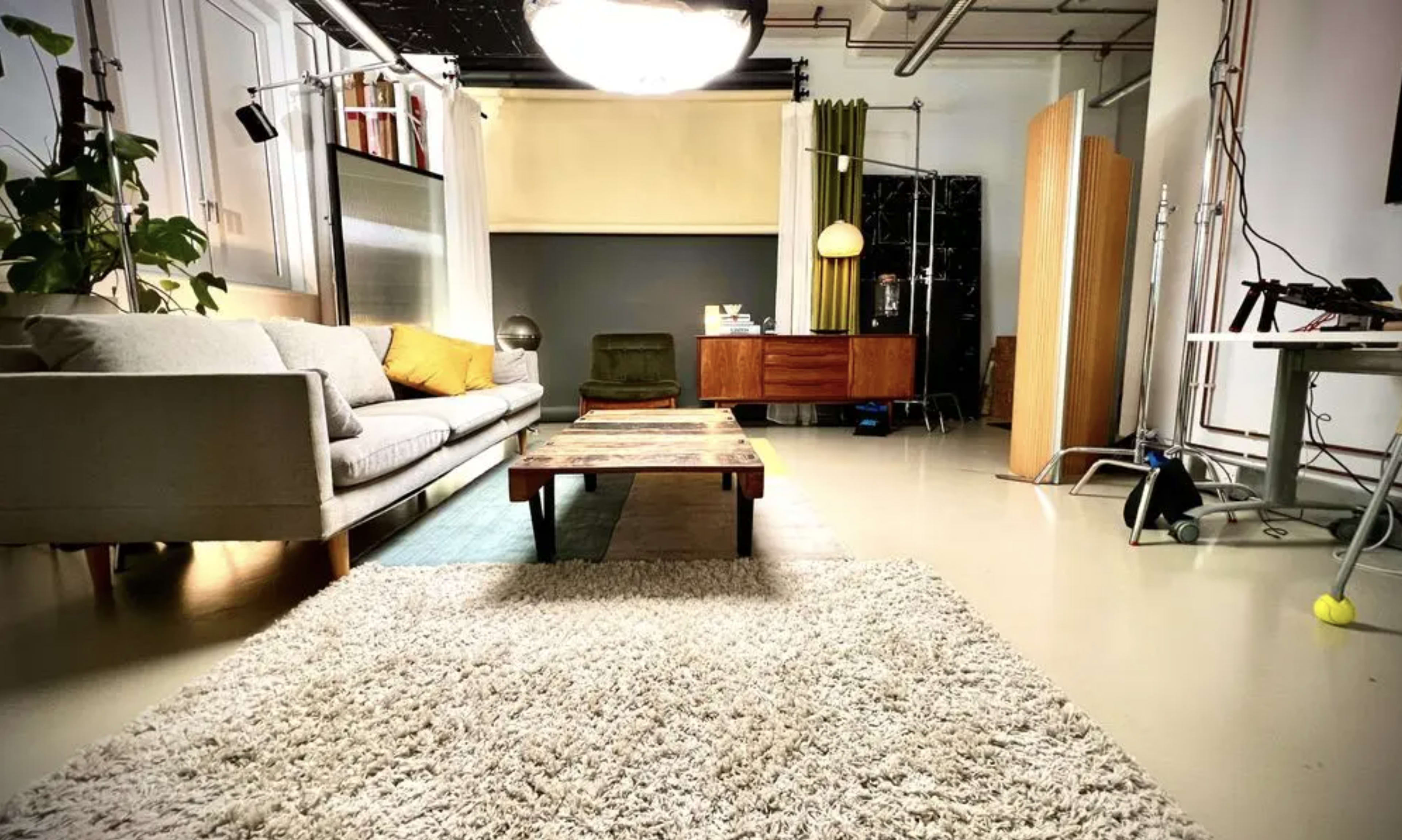 A spacious living area features a gray sofa, a wooden coffee table, and a rug on a light-colored floor, with a collection of furniture and decor in the background.