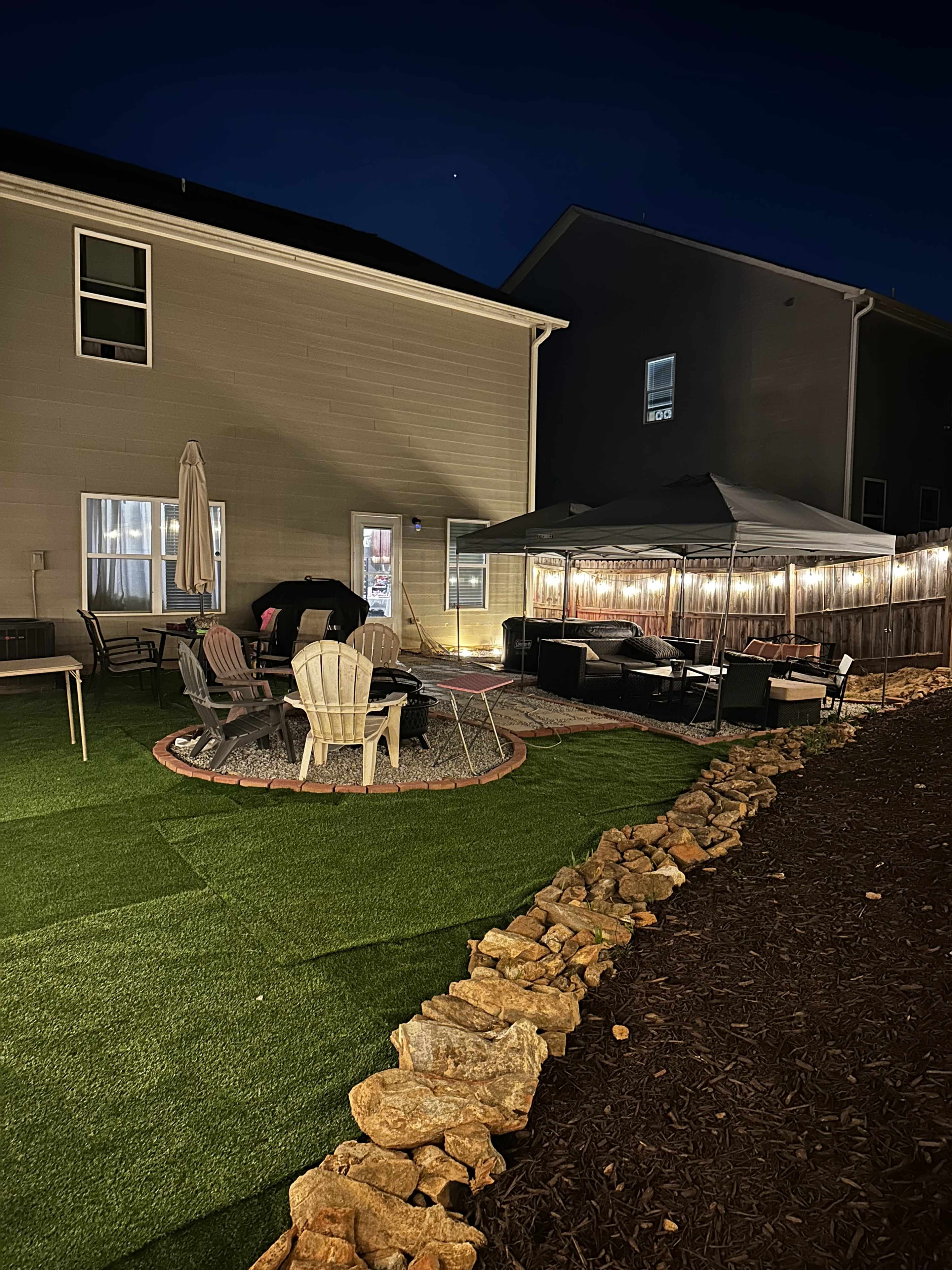 The image shows a backyard at night featuring a circular stone patio with chairs around a fire pit, a canopy tent, and artificial grass areas bordered by a stone wall.