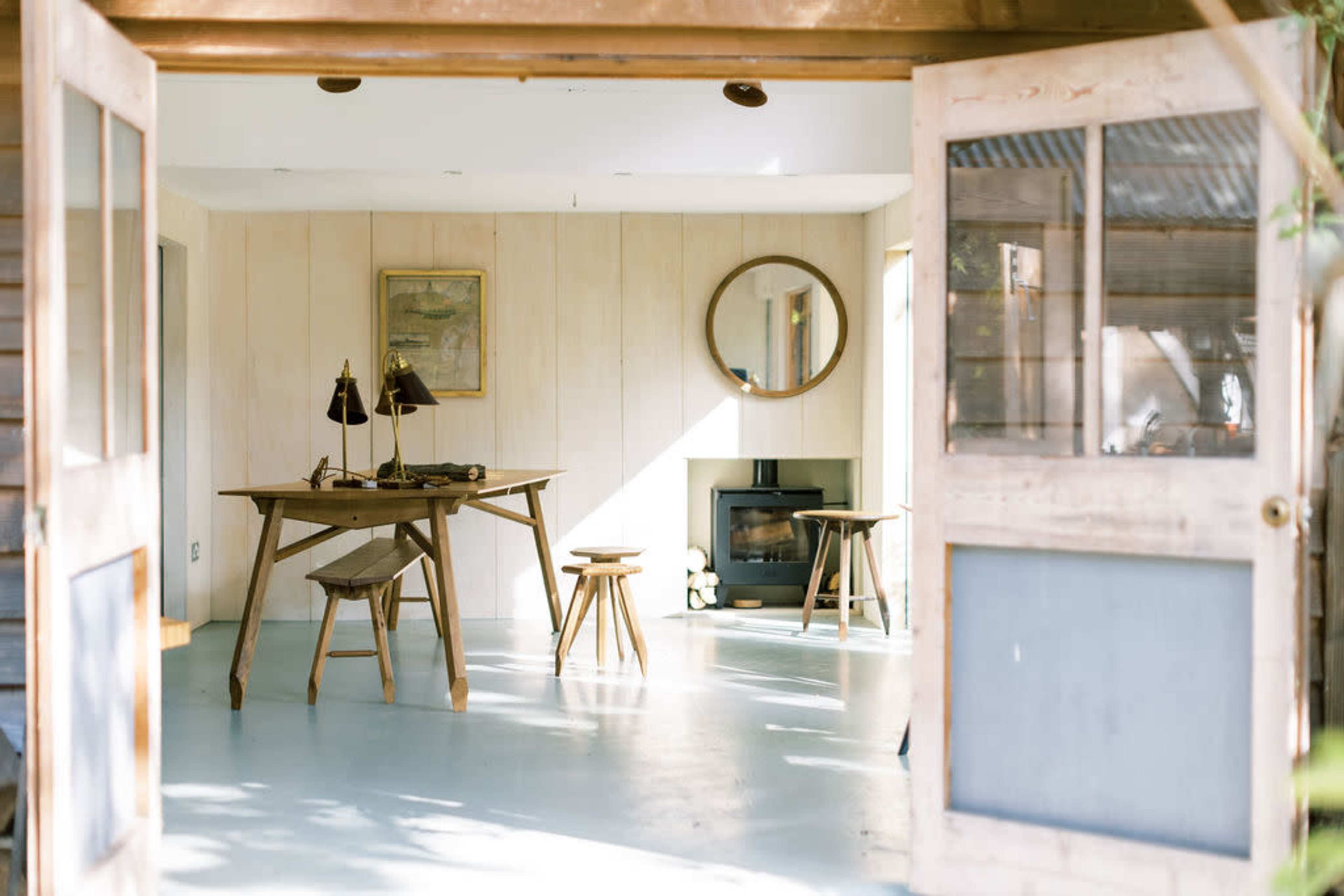 A spacious, minimally decorated room features a wooden table and stools, a round mirror on the wall, and a fireplace.