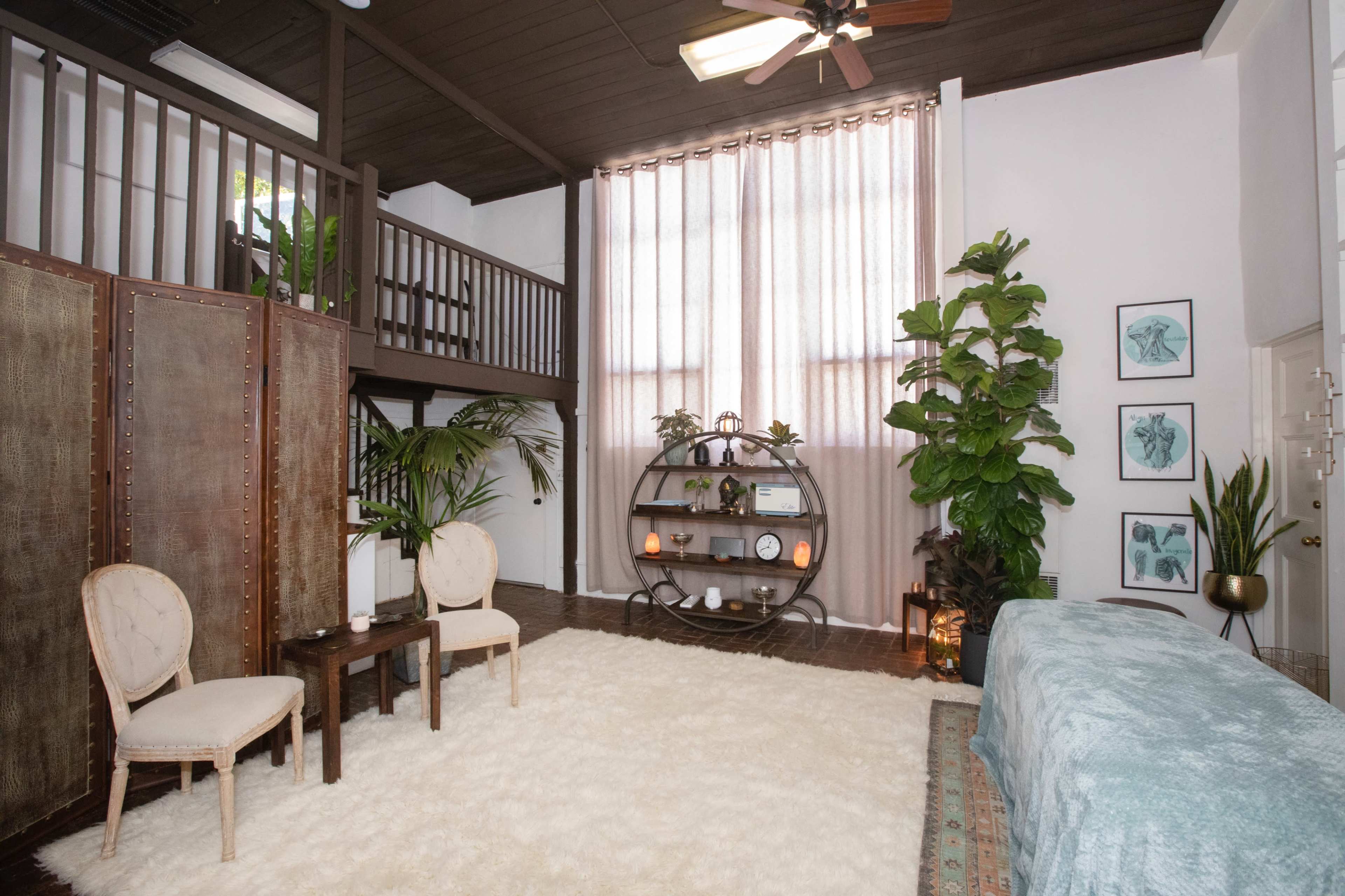 A well-lit room featuring a cozy seating area with two chairs and a small table, a large bed, decorative plants, and a tall window with sheer curtains.
