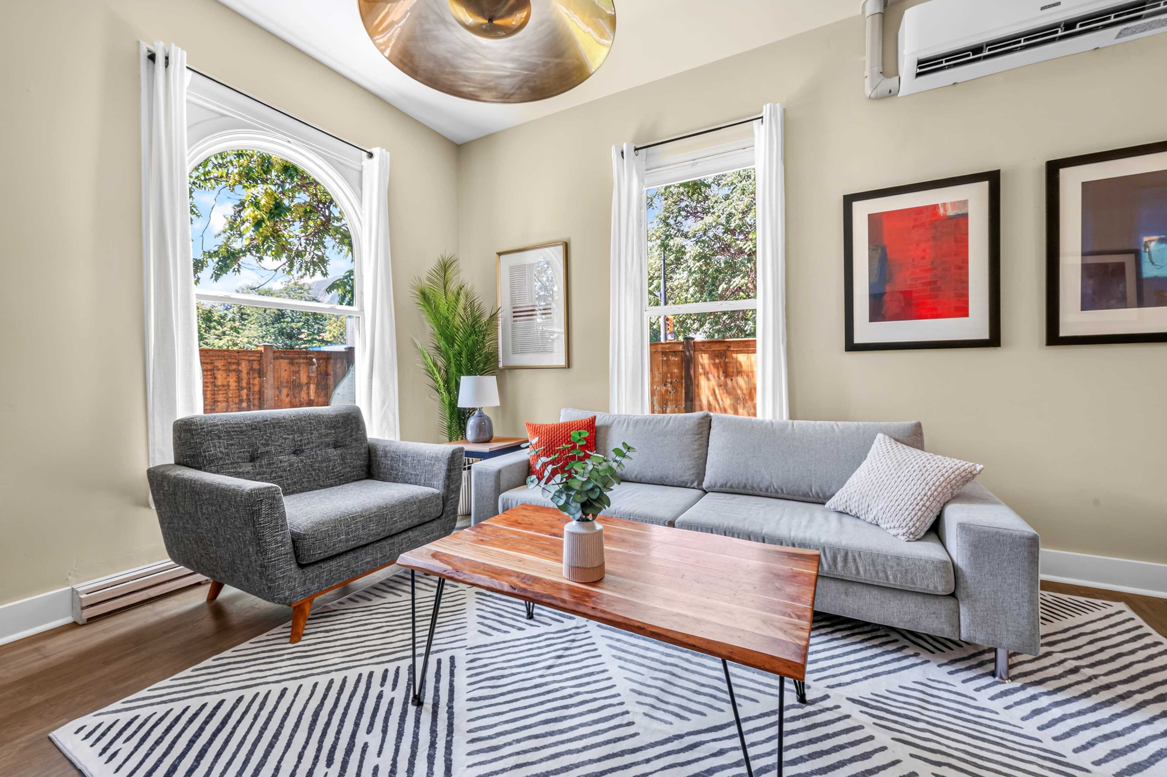 The image shows a modern living room with a gray sofa, an armchair, a wooden coffee table, and large windows framed by white curtains.