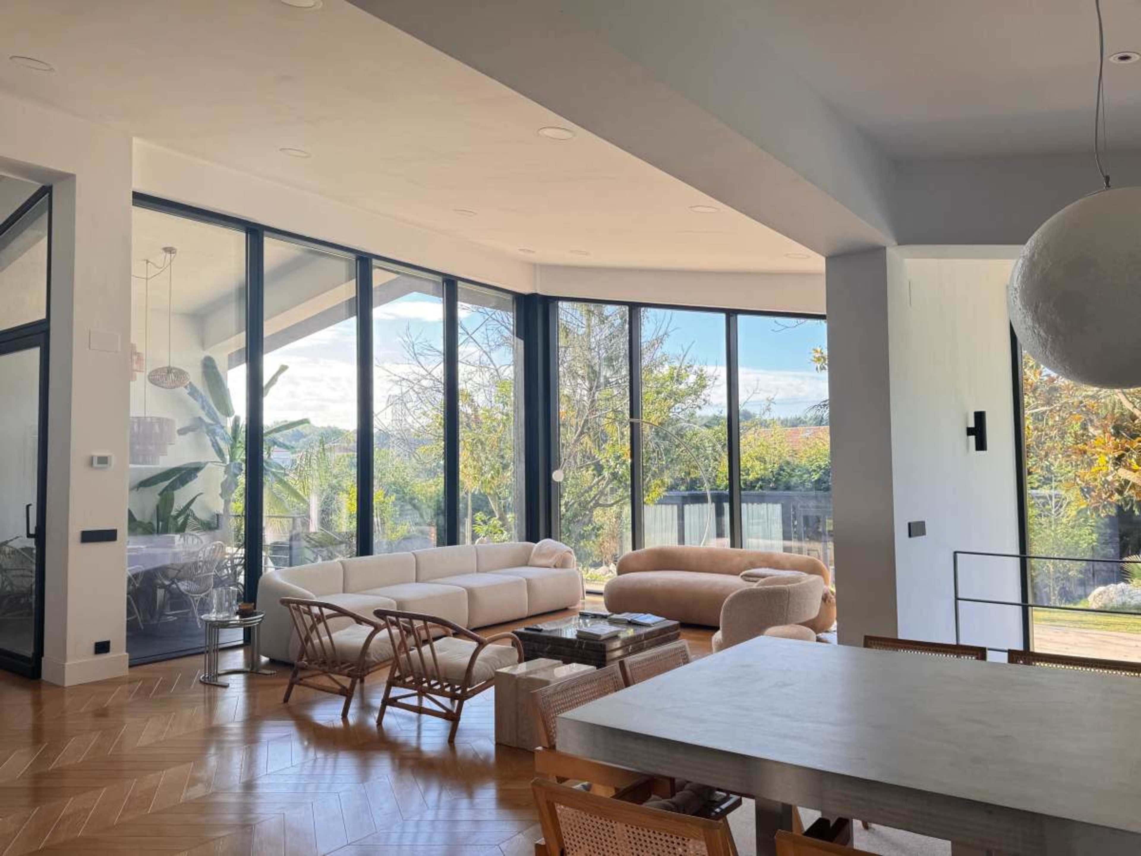 The image shows a modern living space with large windows, stylish furniture, and a dining area, surrounded by greenery.