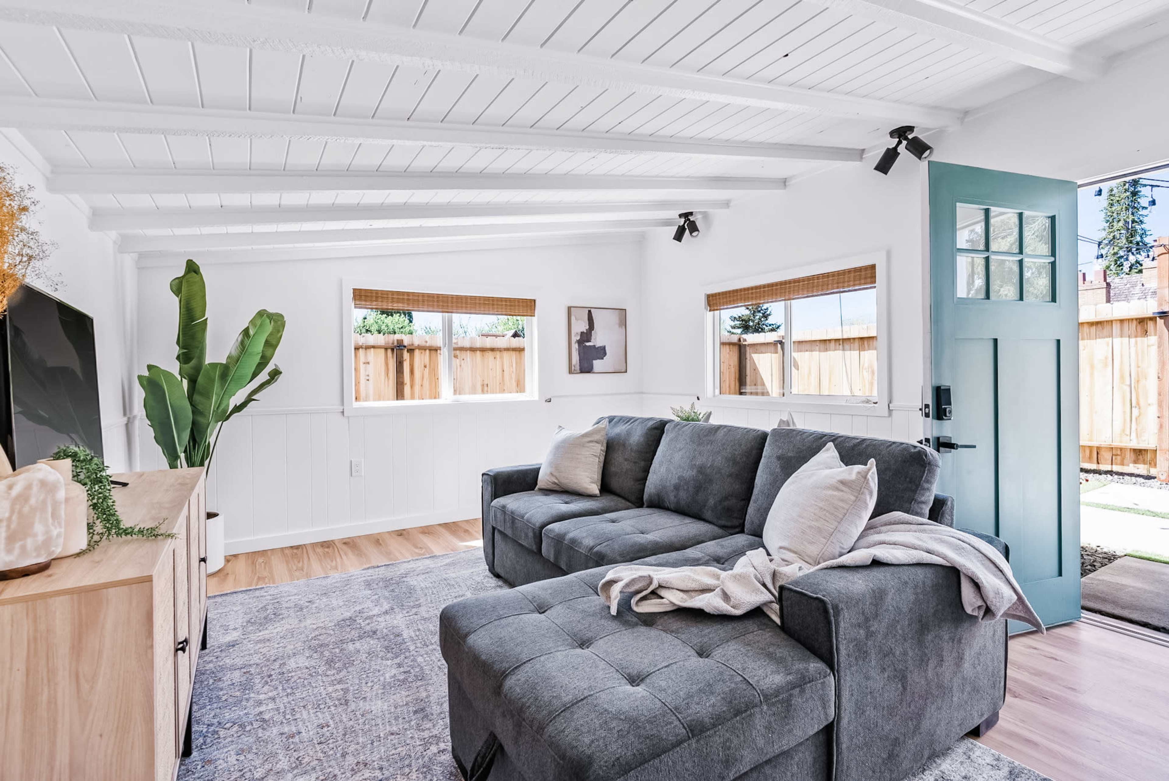 The room features a gray sectional sofa, a light wooden sideboard, and large windows that illuminate the space.