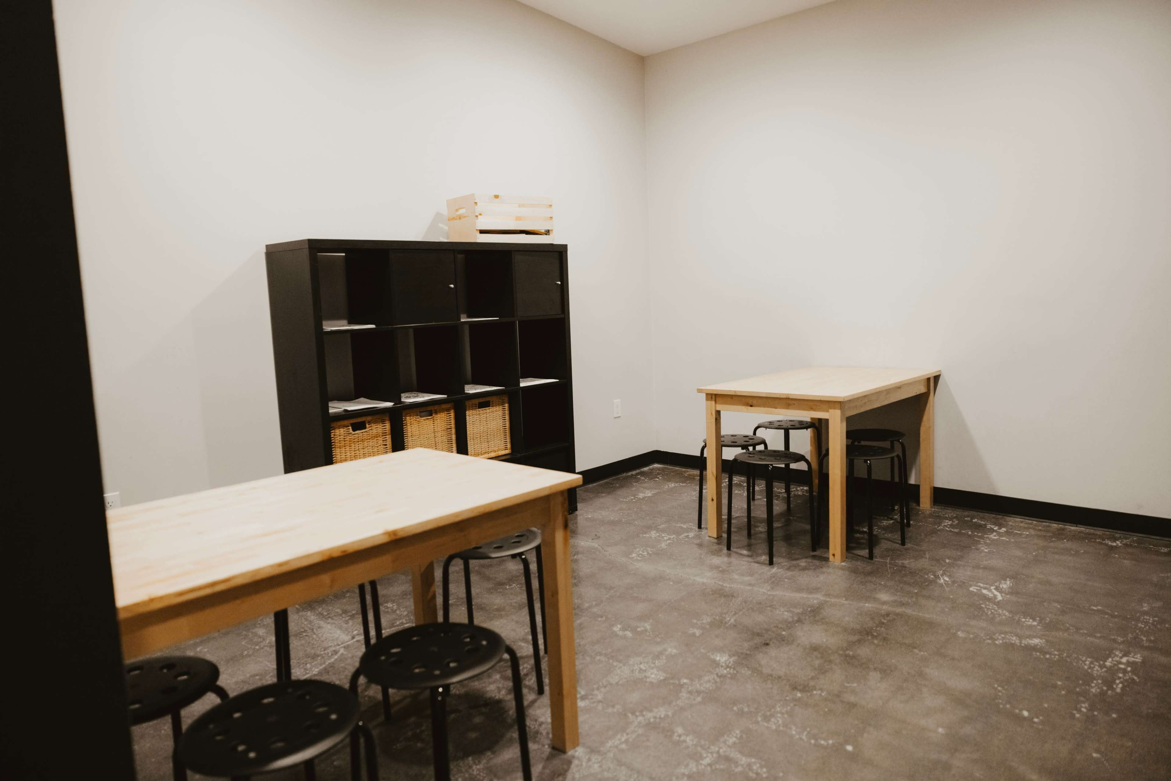 The image shows a simple, empty room with two wooden tables and black stools, alongside a black shelving unit containing storage baskets.