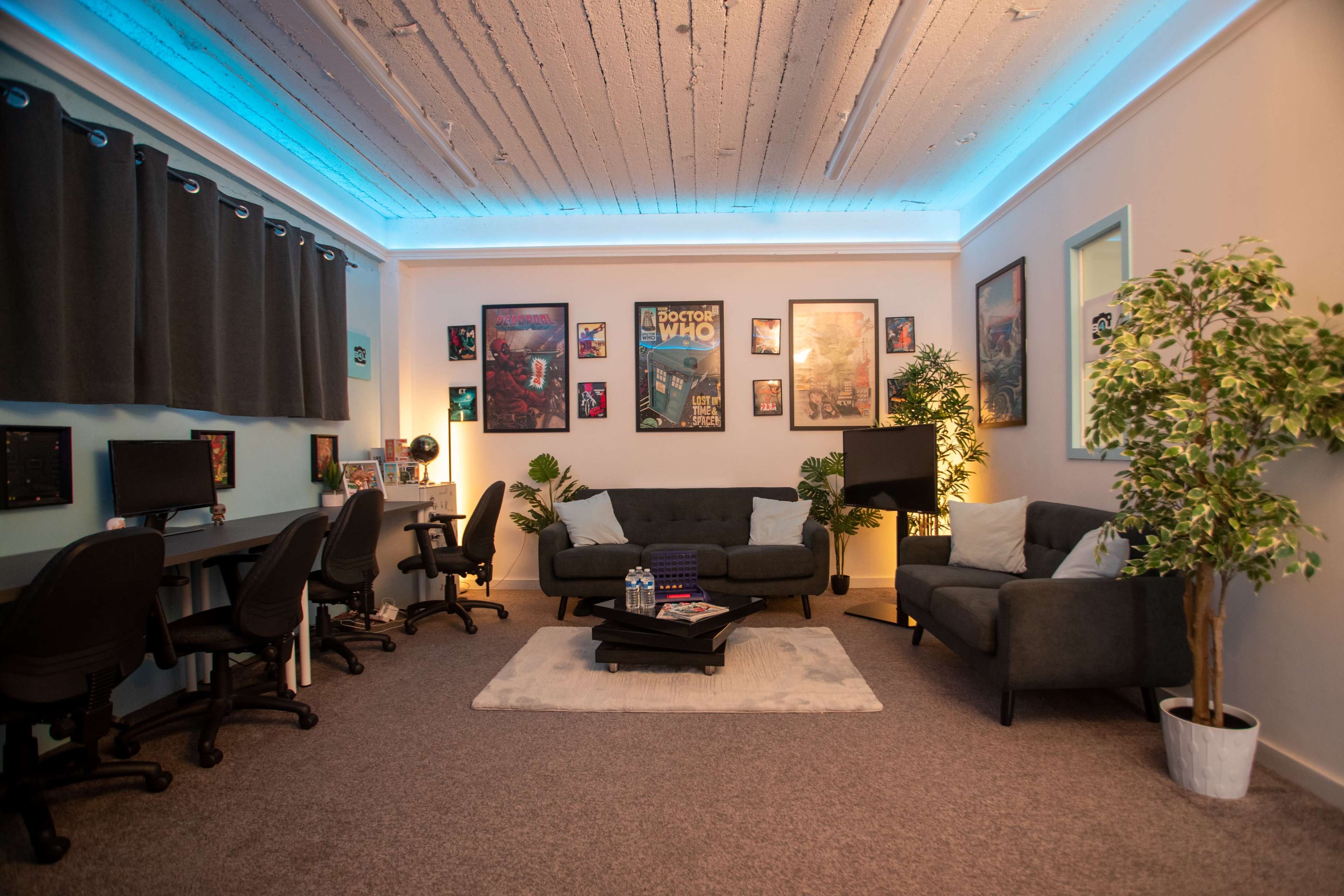 A modern office space with four ergonomic chairs, two couches, framed posters on the walls, and plants, all under soft blue lighting.