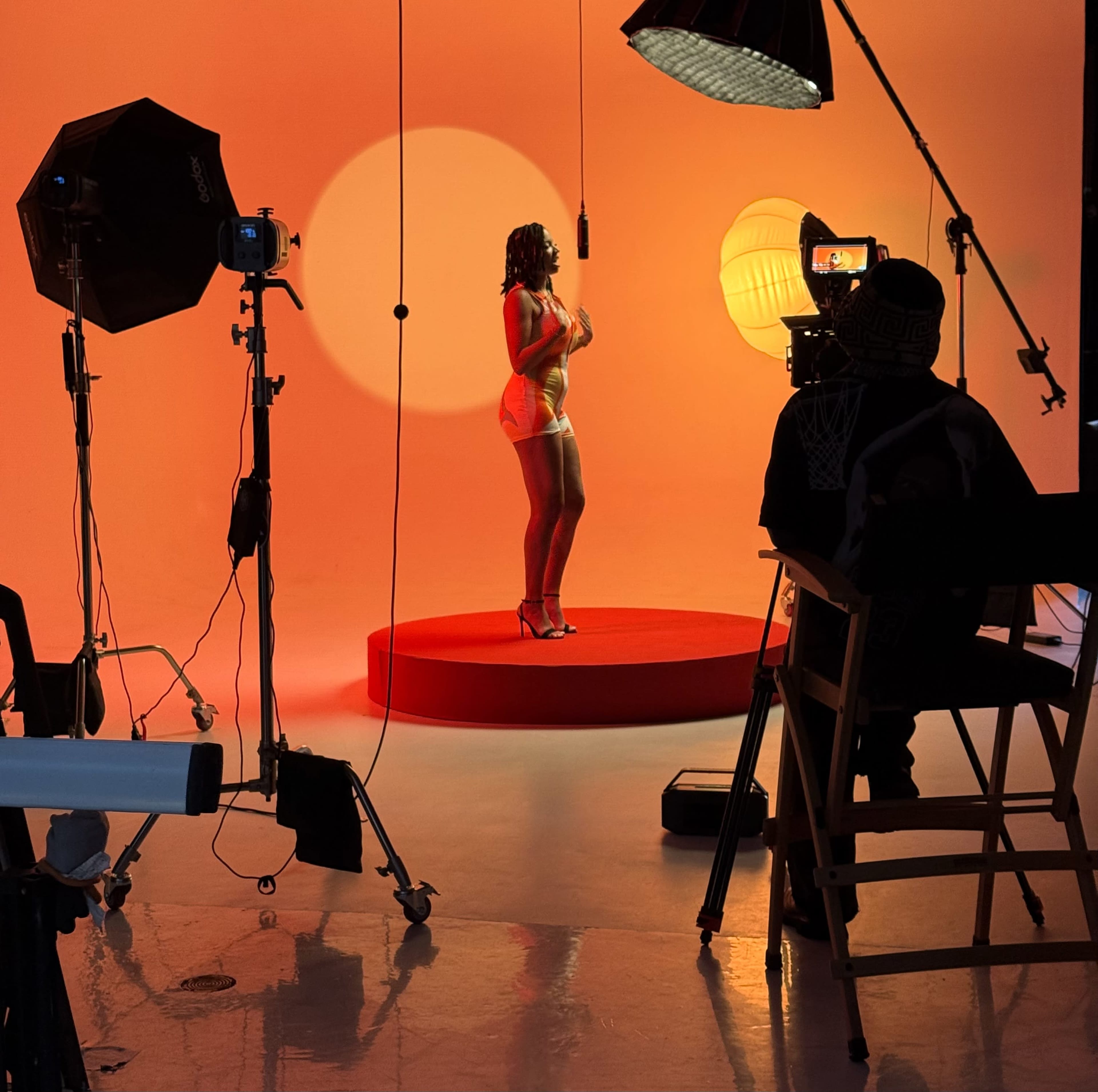 A model stands on a circular platform in front of an orange backdrop, surrounded by photography equipment and lighting.