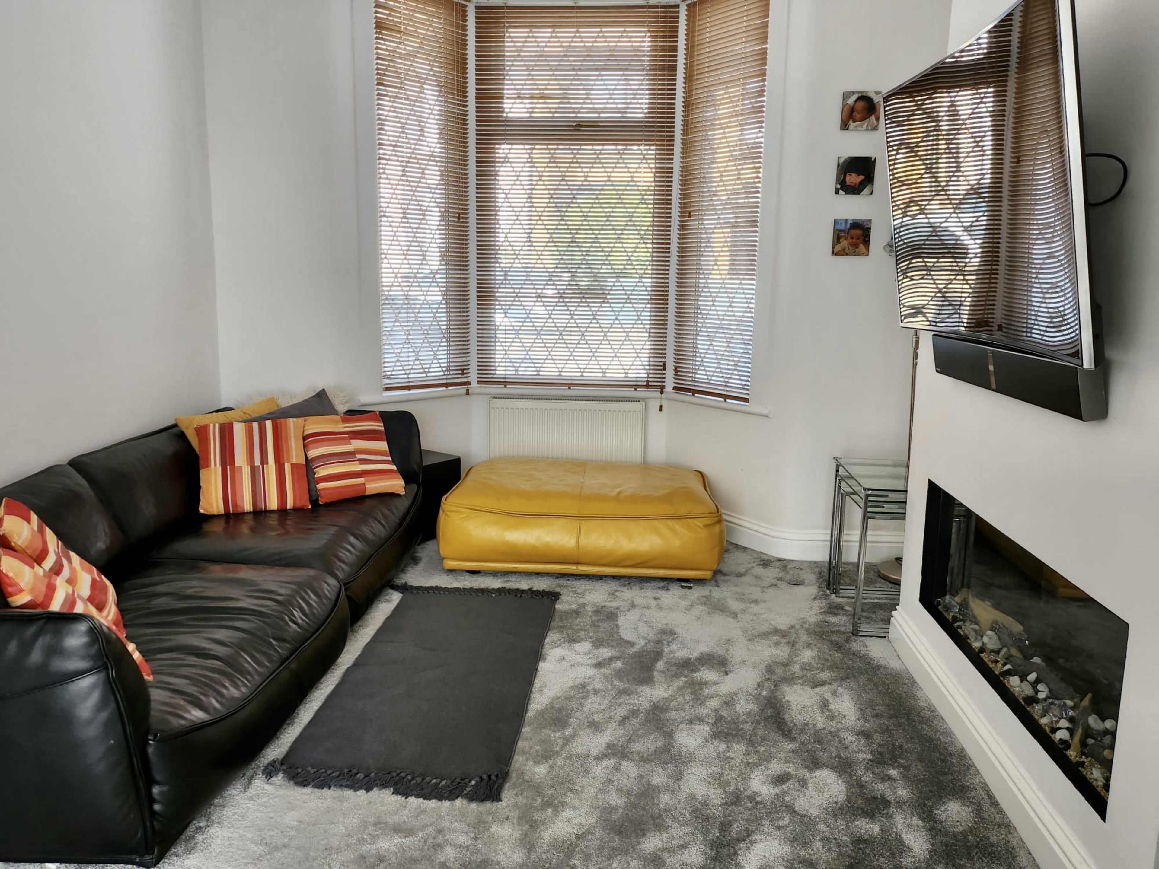 The image shows a living room featuring a black leather sofa with striped cushions, a yellow ottoman, a television mounted on the wall, and a fireplace below it, with large windows covered by wooden blinds.