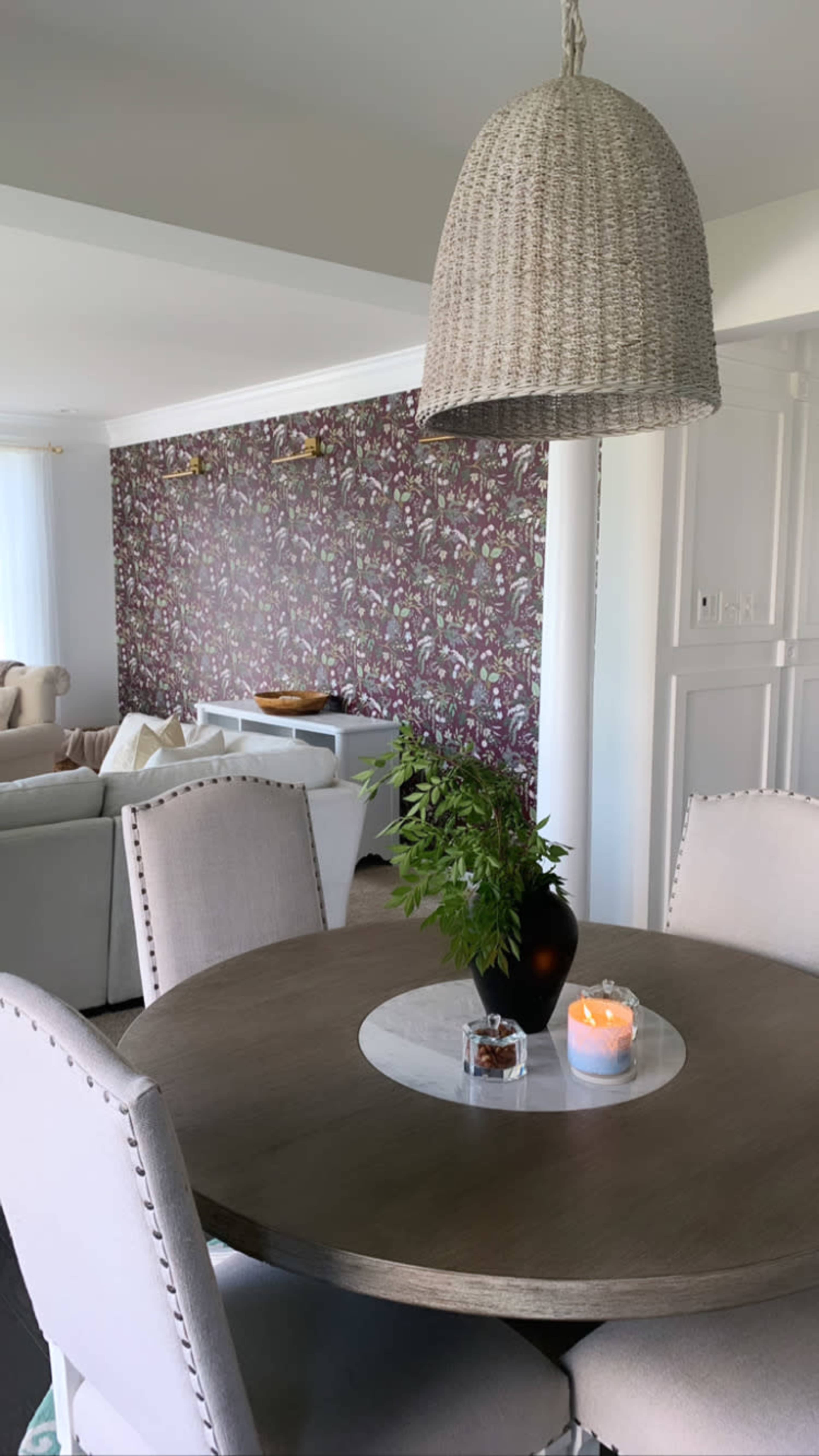 A round dining table with a vase of greenery and a lit candle is situated under a woven pendant light, with a patterned wallpaper in the background and seating areas visible.