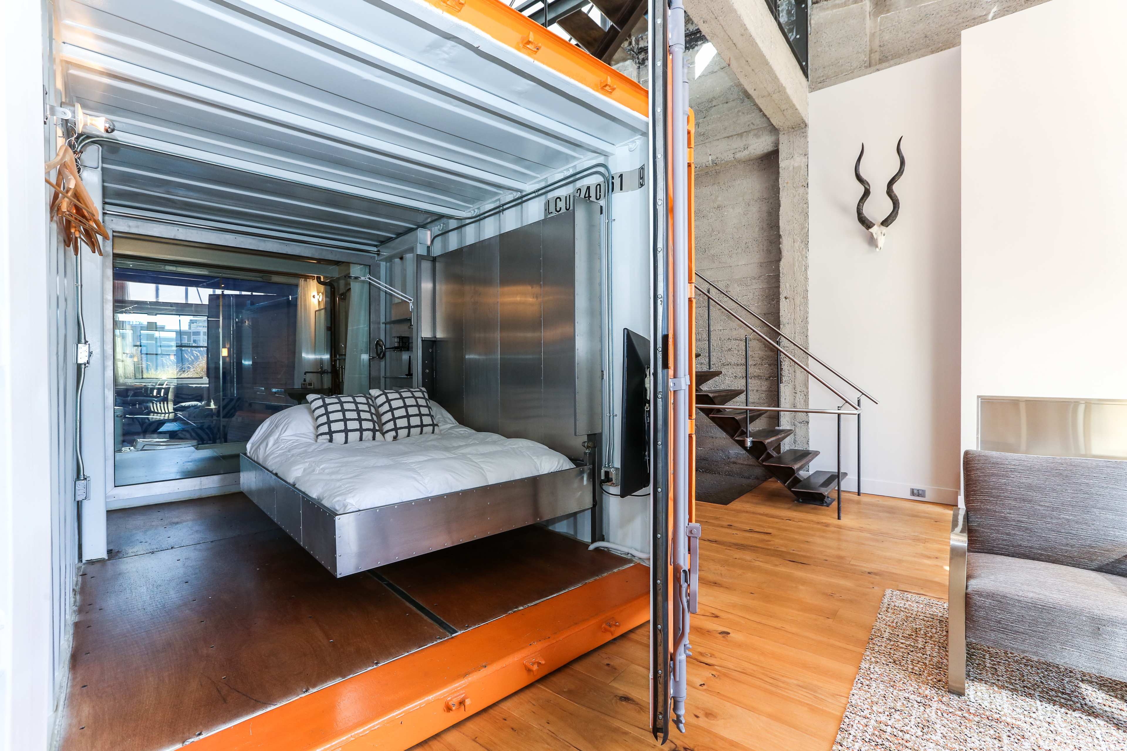 The image shows a modern bedroom designed within a shipping container, featuring a bed with white linens and a staircase leading to a lofted area.