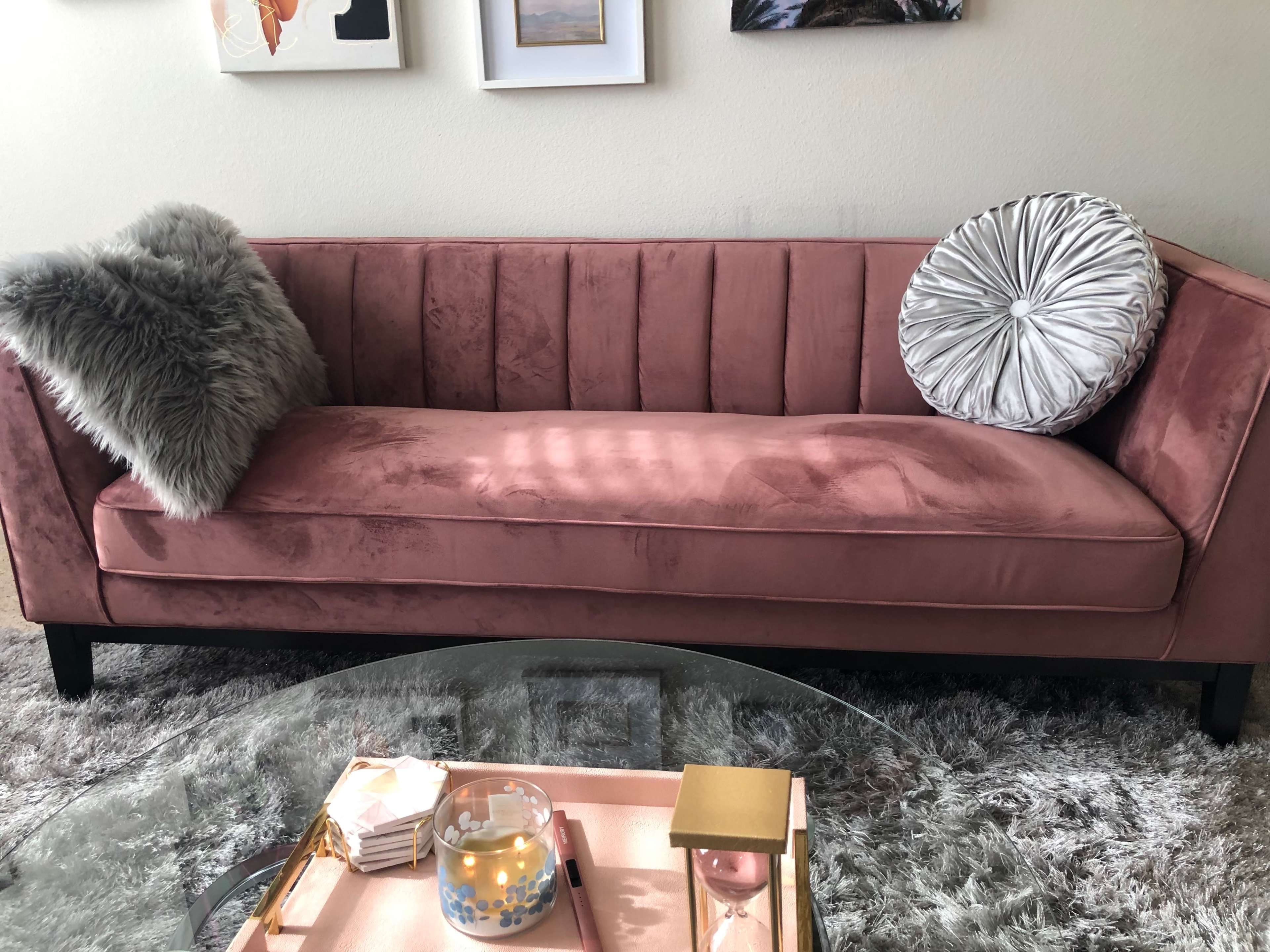 A pink velvet sofa with a textured cushion and a decorative pillow is positioned near a glass coffee table on a plush rug.