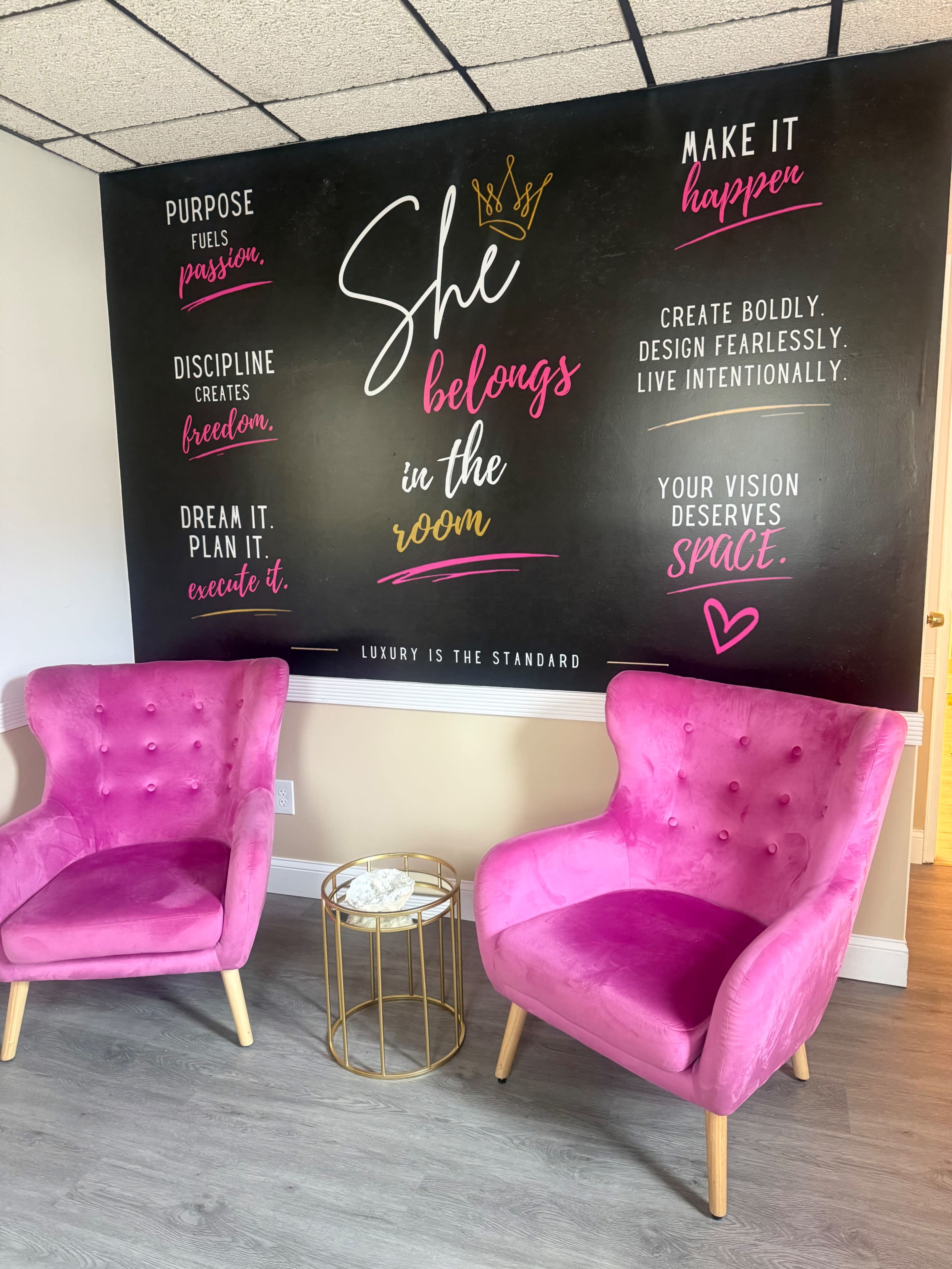 The image shows a stylish room with two pink velvet chairs facing a large wall mural that features motivational quotes in various colors.