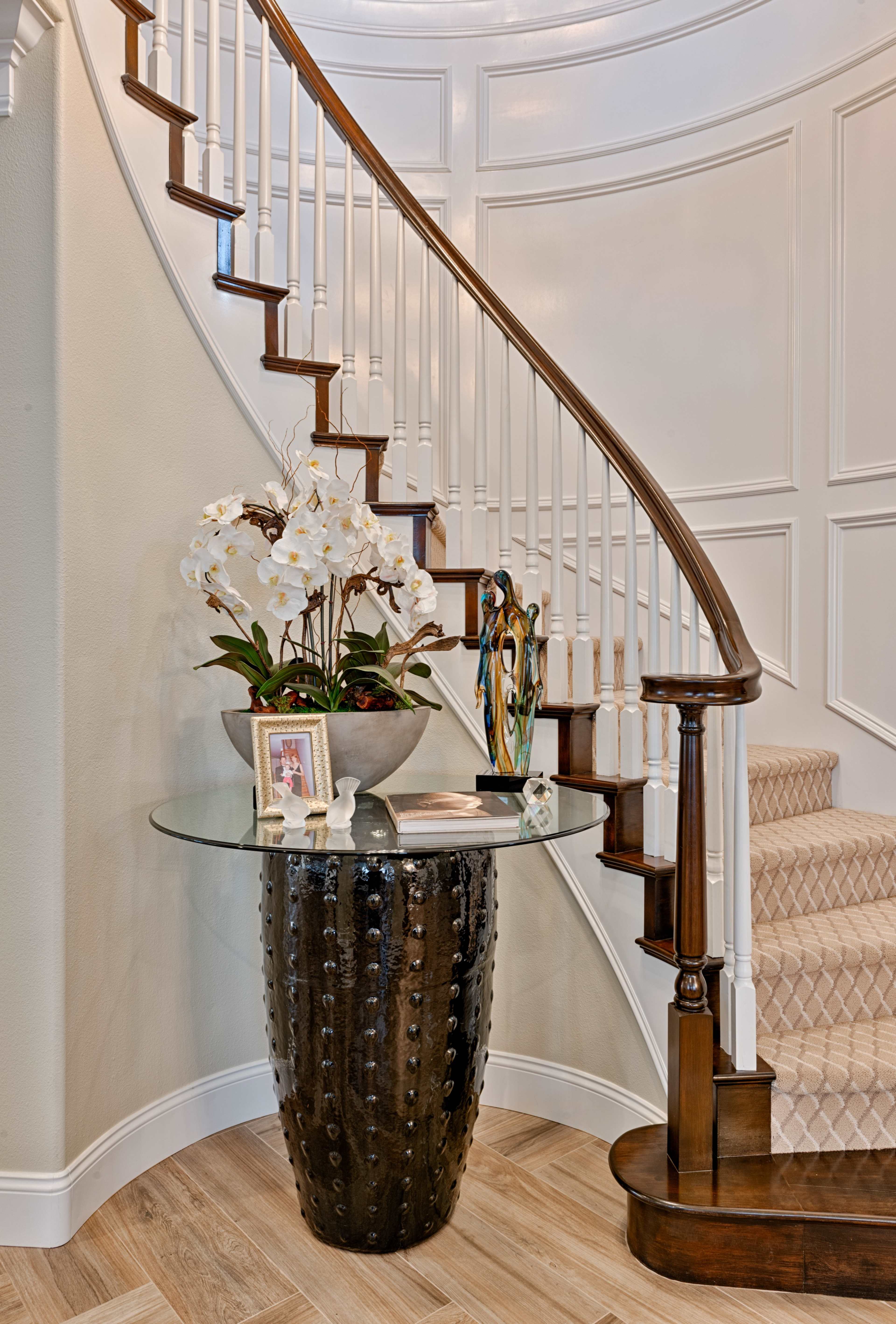 A curved staircase with wooden railings surrounds a decorative table featuring a glass top and a floral arrangement, positioned in a well-lit foyer.