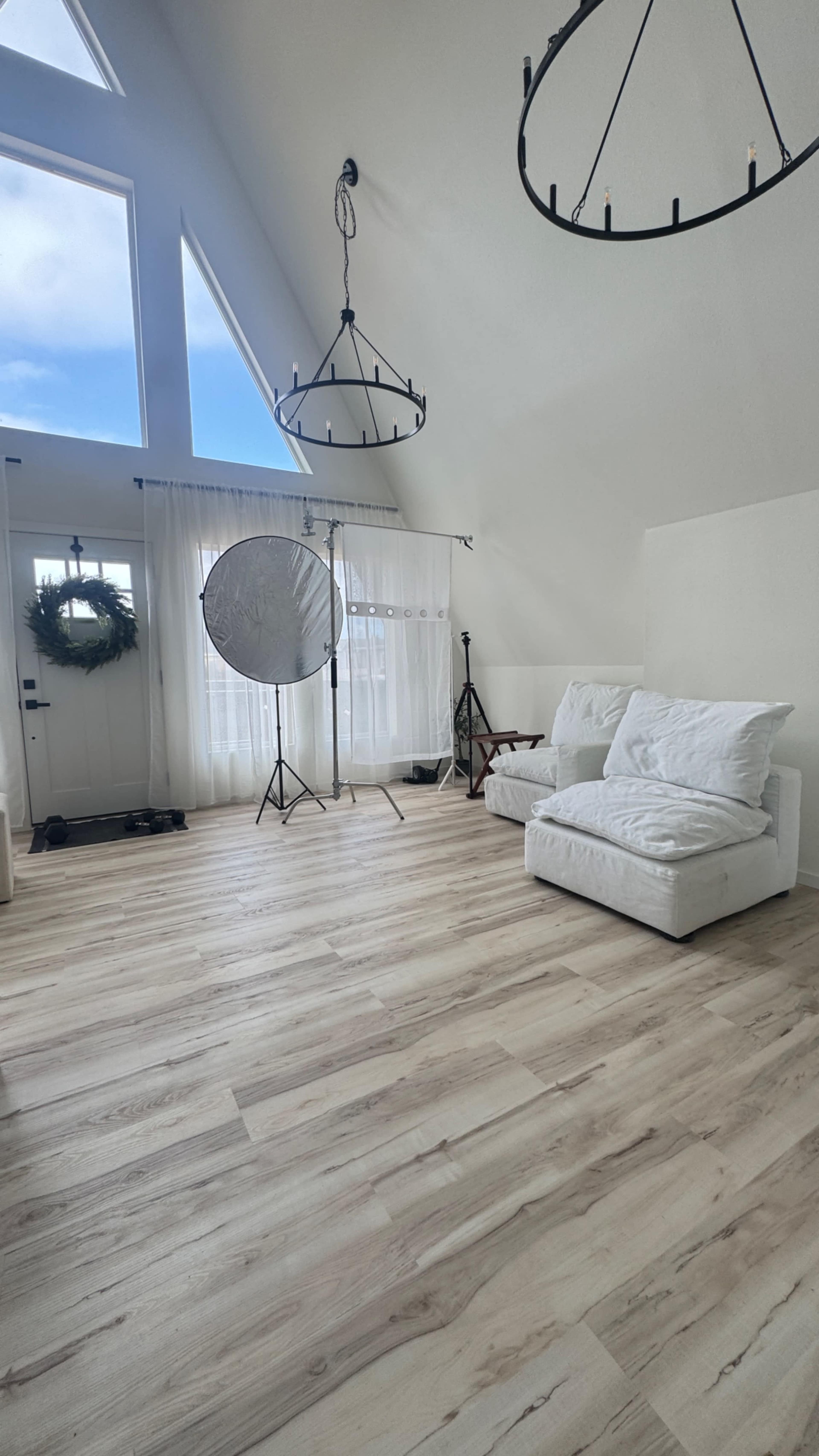 The image features a bright, spacious room with high ceilings, large windows, and minimalistic furniture, including two white sofas and a set of photography equipment.