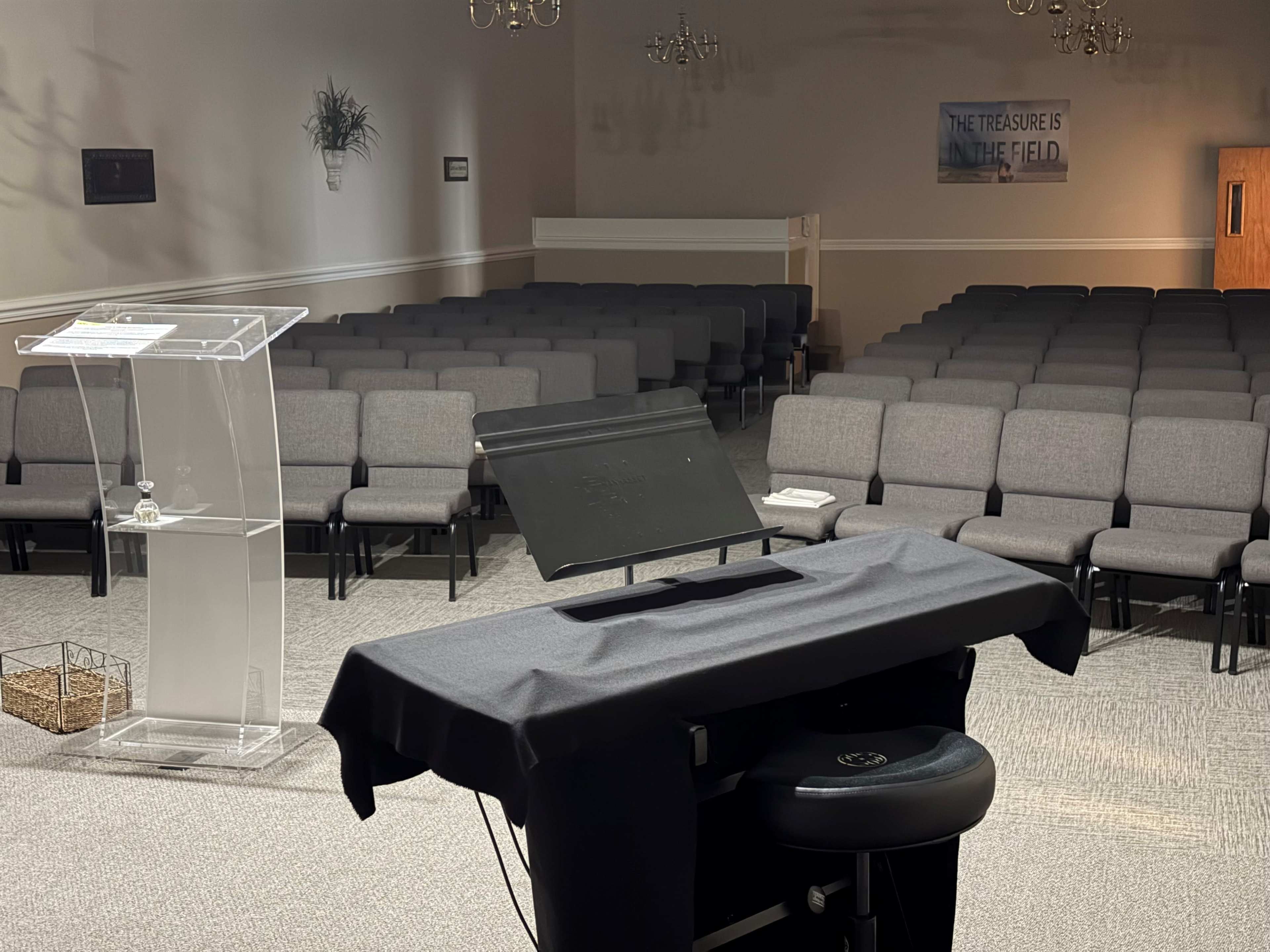 The image shows a sparsely decorated room with rows of gray chairs arranged for an event, featuring a clear podium and a music stand at the front.