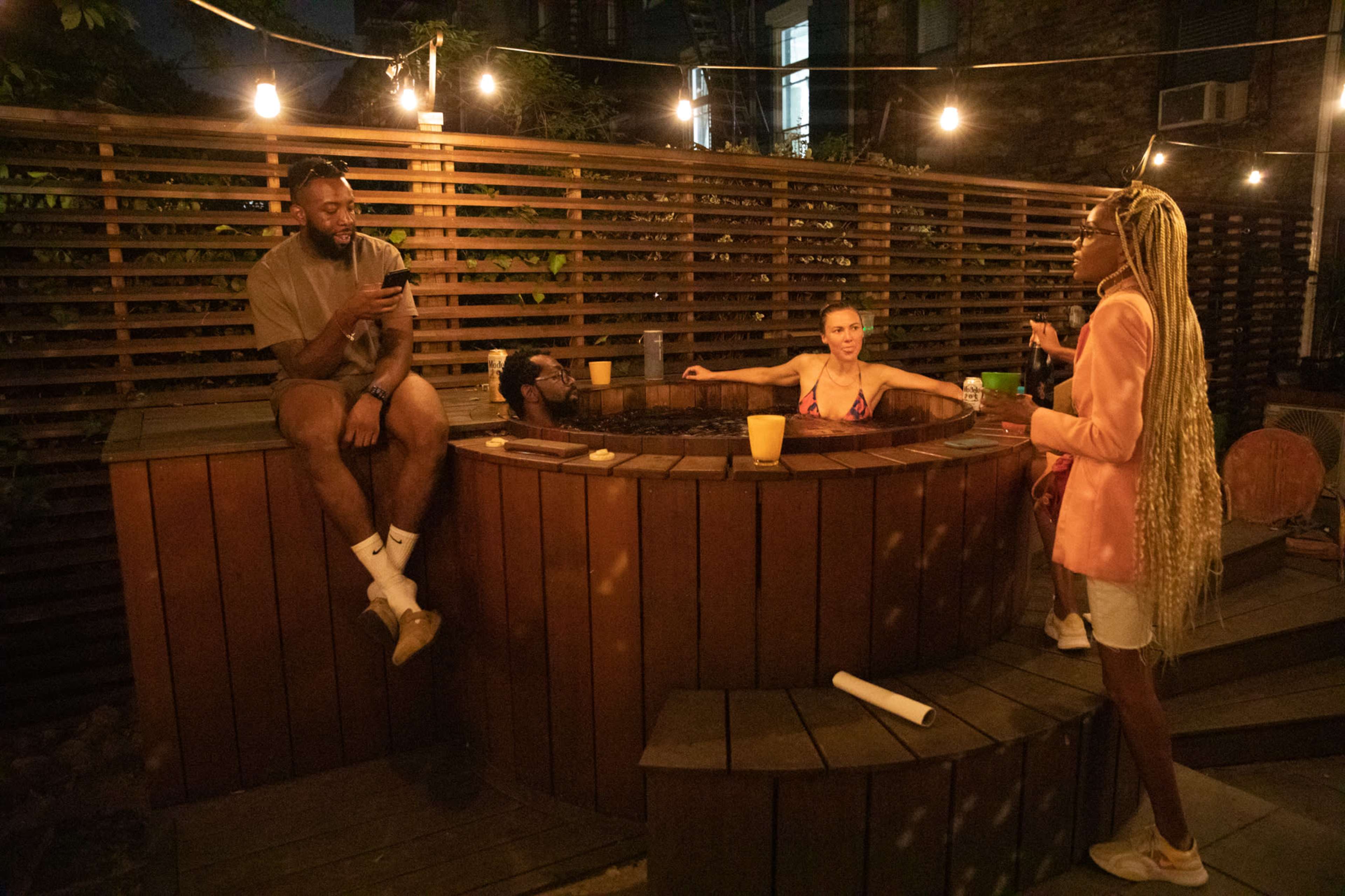 A group of four people socialize around a wooden hot tub in a dimly lit outdoor setting, with string lights overhead.