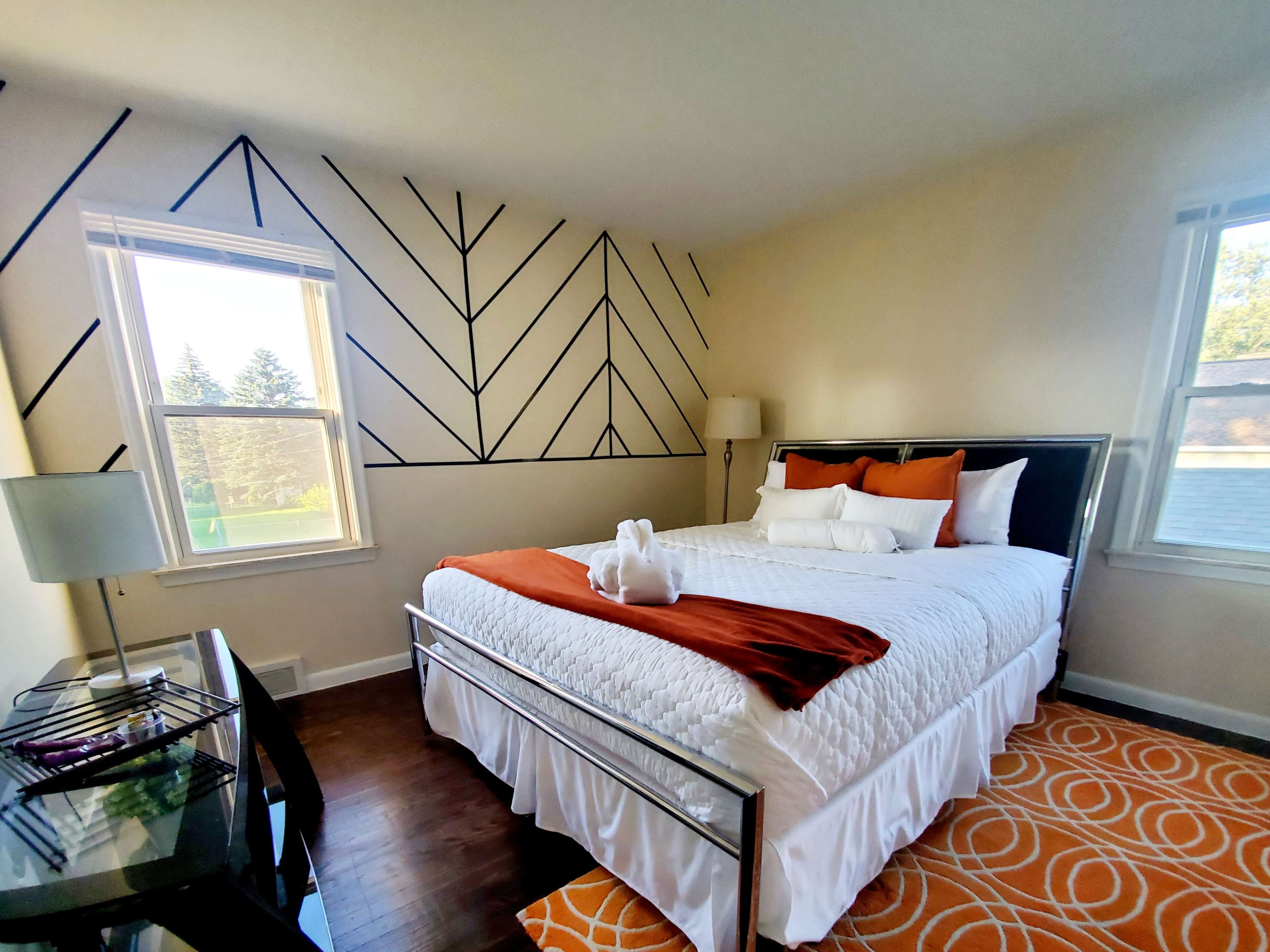 The room features a double bed with white and orange bedding, a patterned area rug, and a geometric wall design, along with large windows letting in natural light.