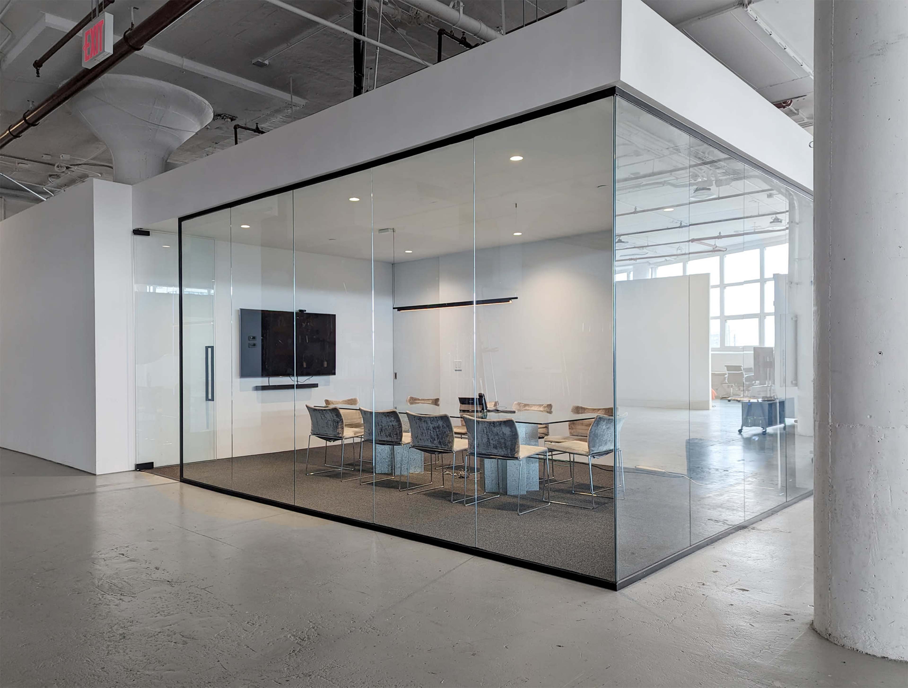 The image shows a modern conference room enclosed in clear glass walls, featuring a large table with chairs and a wall-mounted screen.