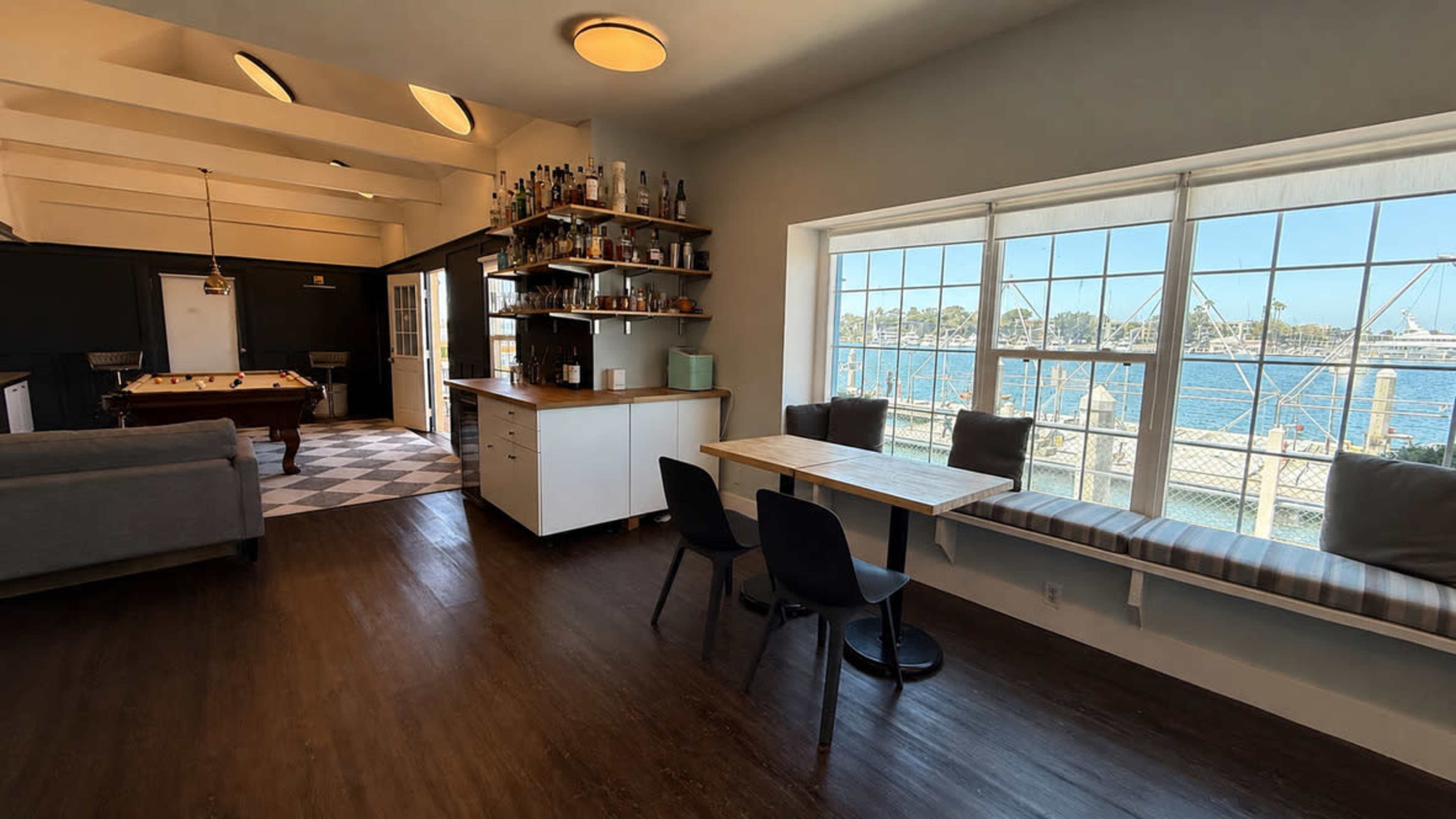 The image shows a bright, open room with a dining table and chairs next to a large window overlooking a waterfront, along with a bar shelving unit and a pool table visible in the adjacent space.