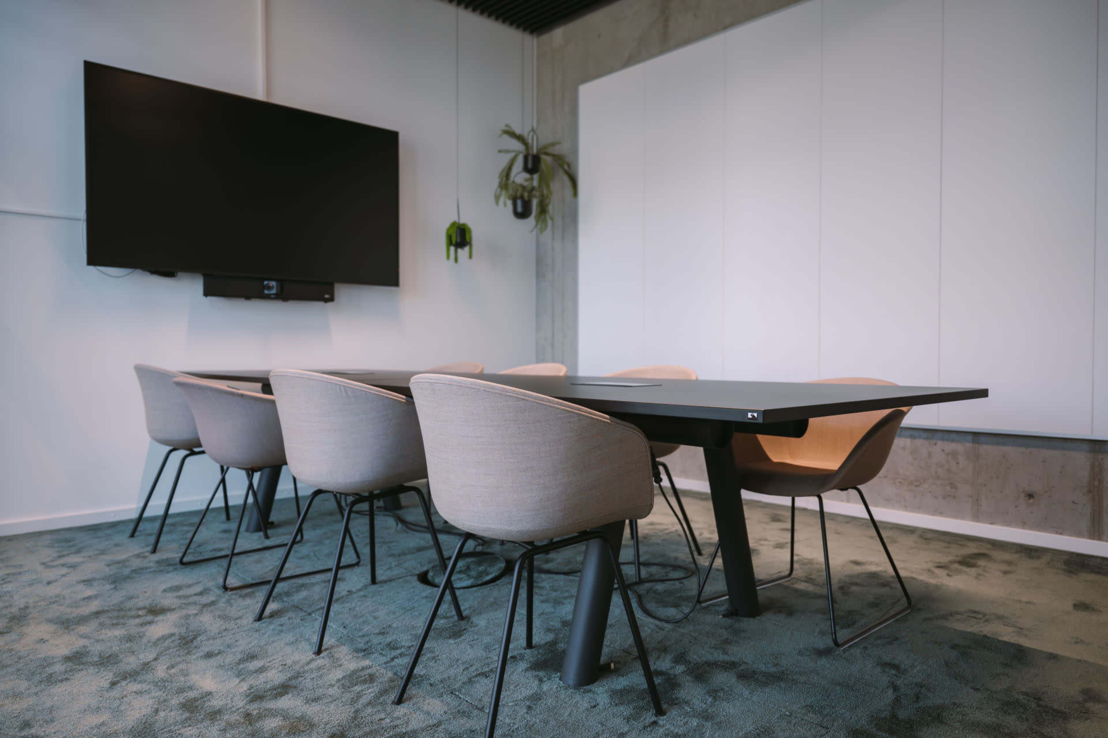 A modern conference room features a large black table surrounded by several beige chairs, with a television mounted on the wall.