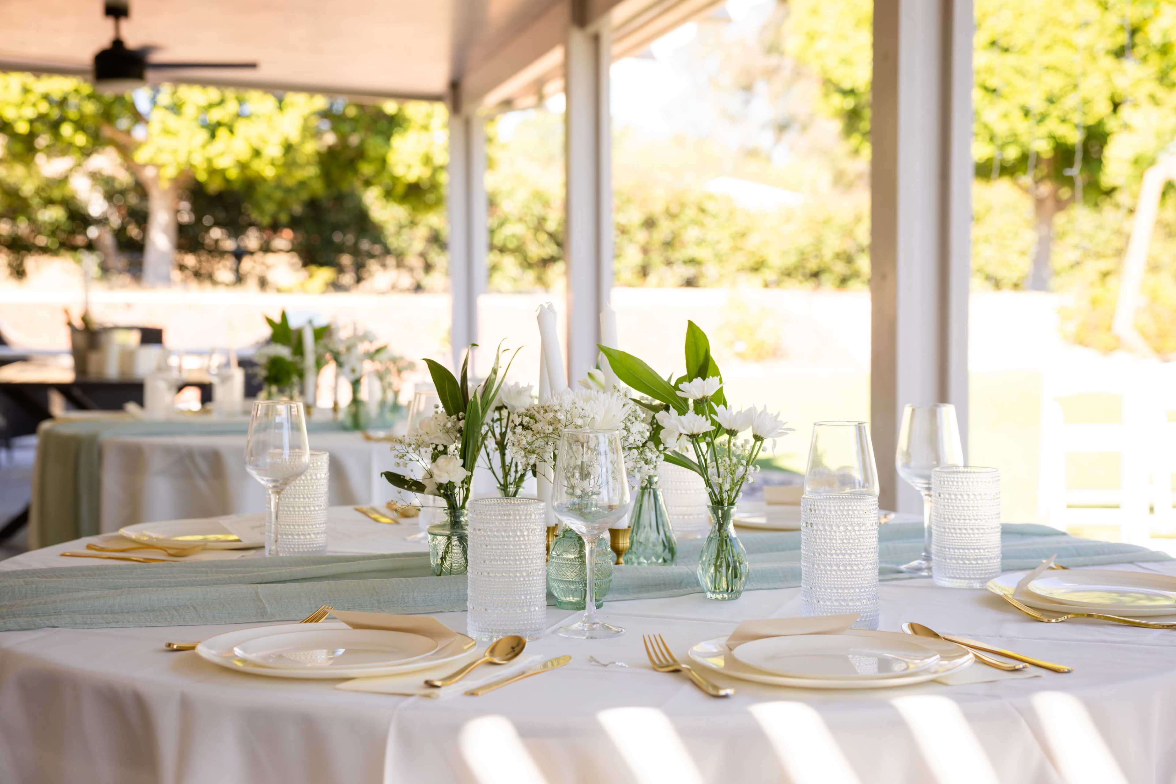 A dining setup features elegantly arranged tables with white tablecloths, glassware, plates, and floral centerpieces under a covered patio.