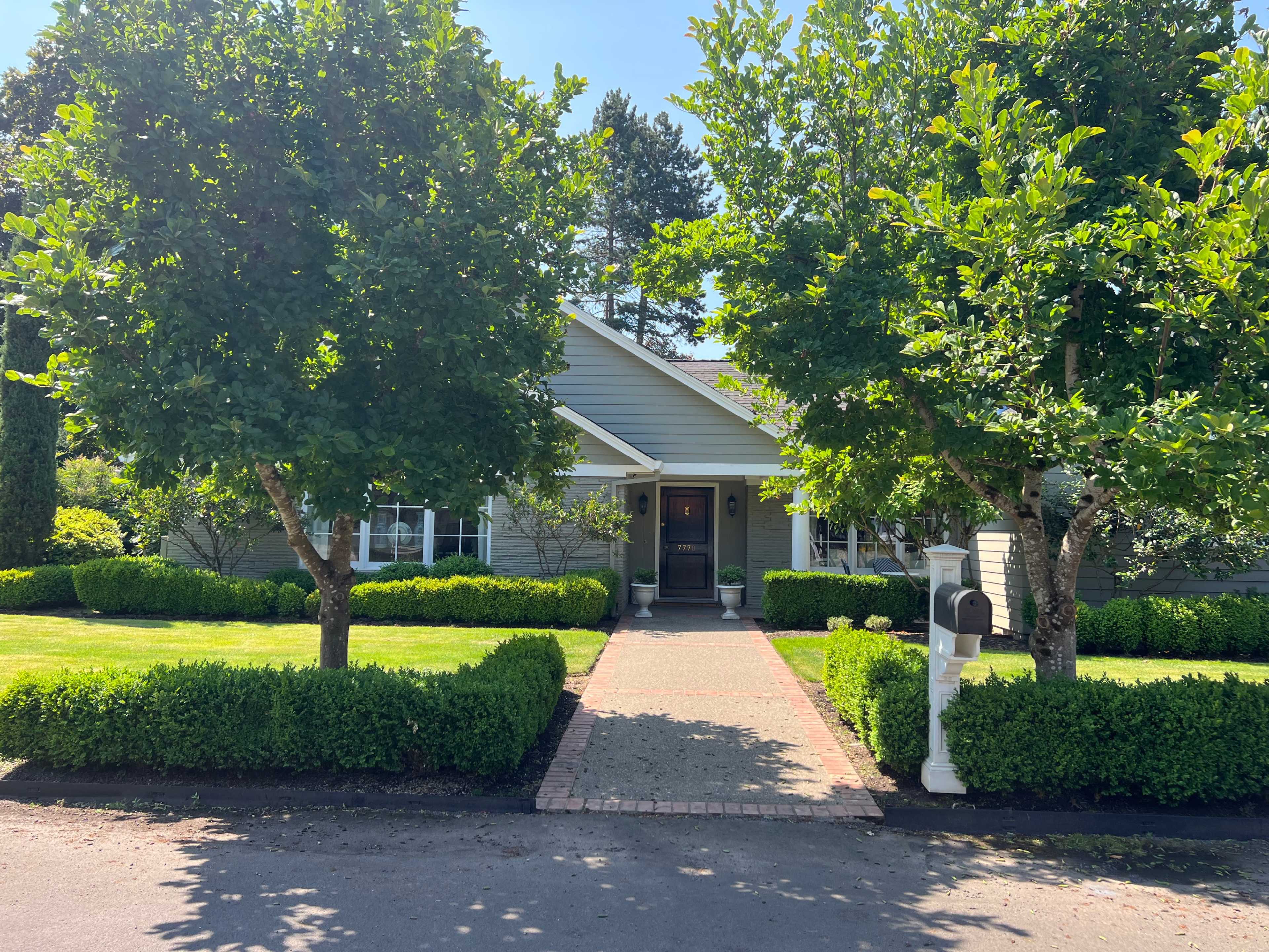 A gray house with a front walkway lined by neatly trimmed hedges and trees leads to a central entrance.
