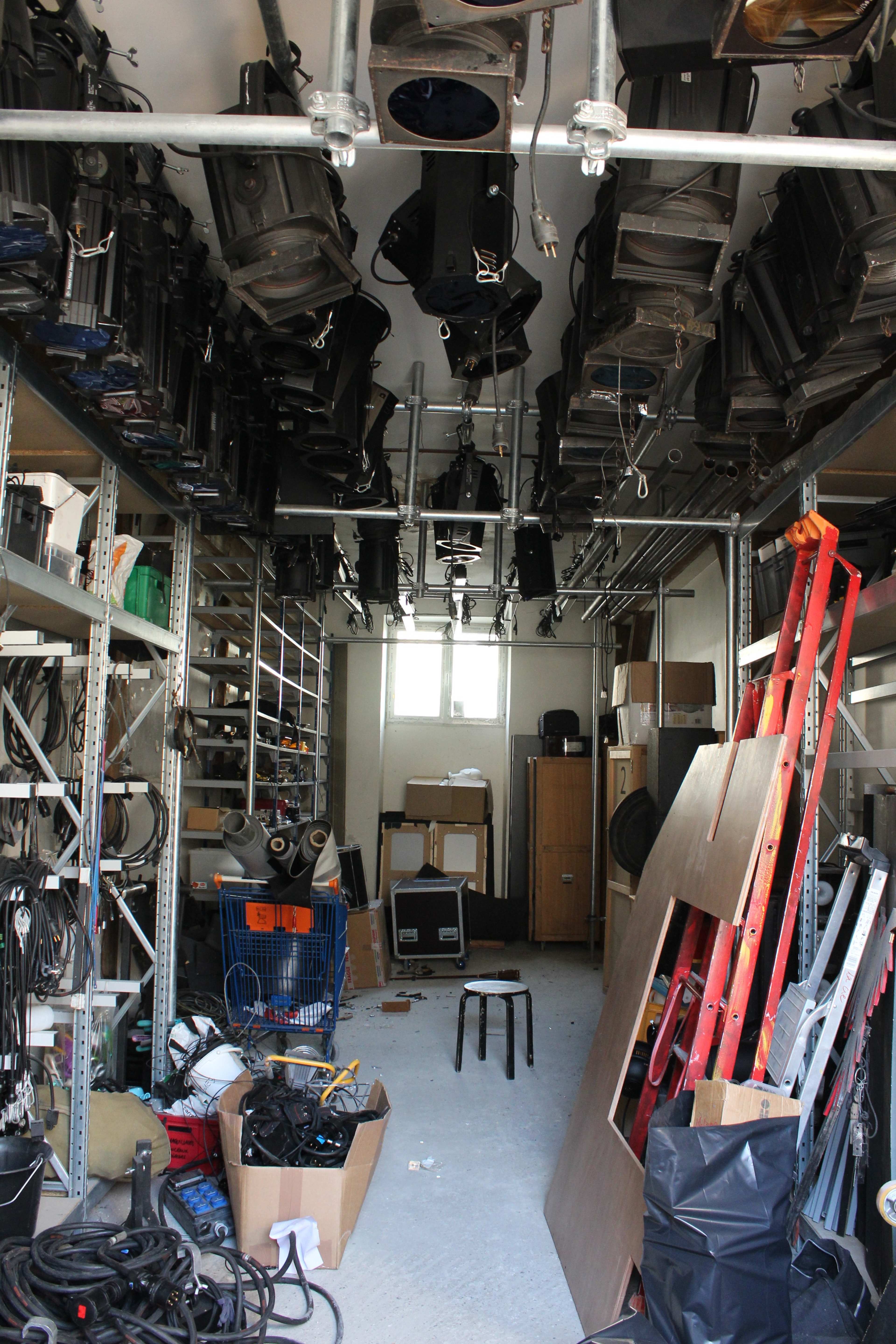 A cluttered storage room filled with lighting equipment, tools, and various materials organized on metal shelving and scattered on the floor.