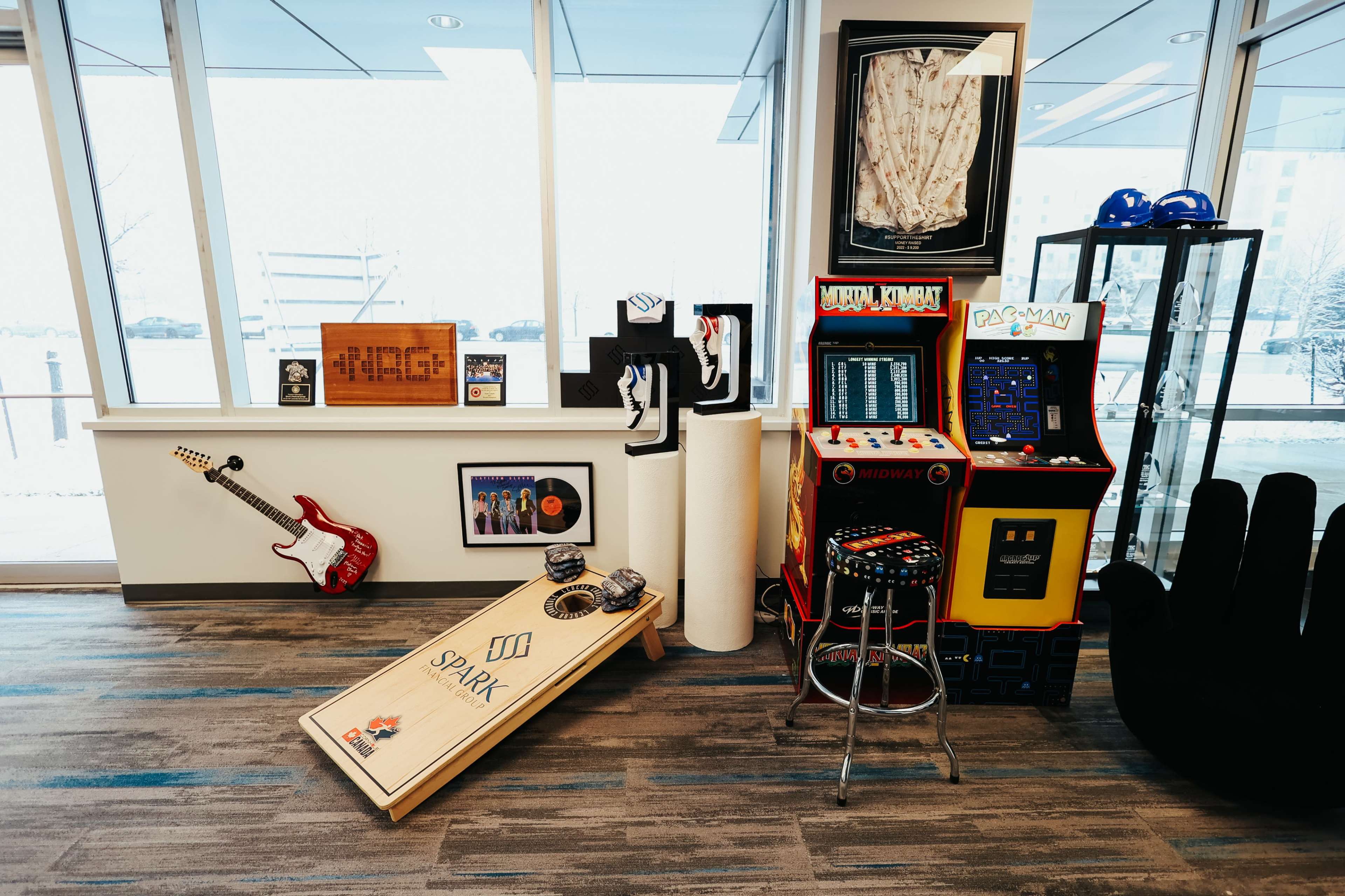 The scene features a playful office space with arcade machines, a cornhole game, and various framed artworks and memorabilia displayed on the walls and furniture.