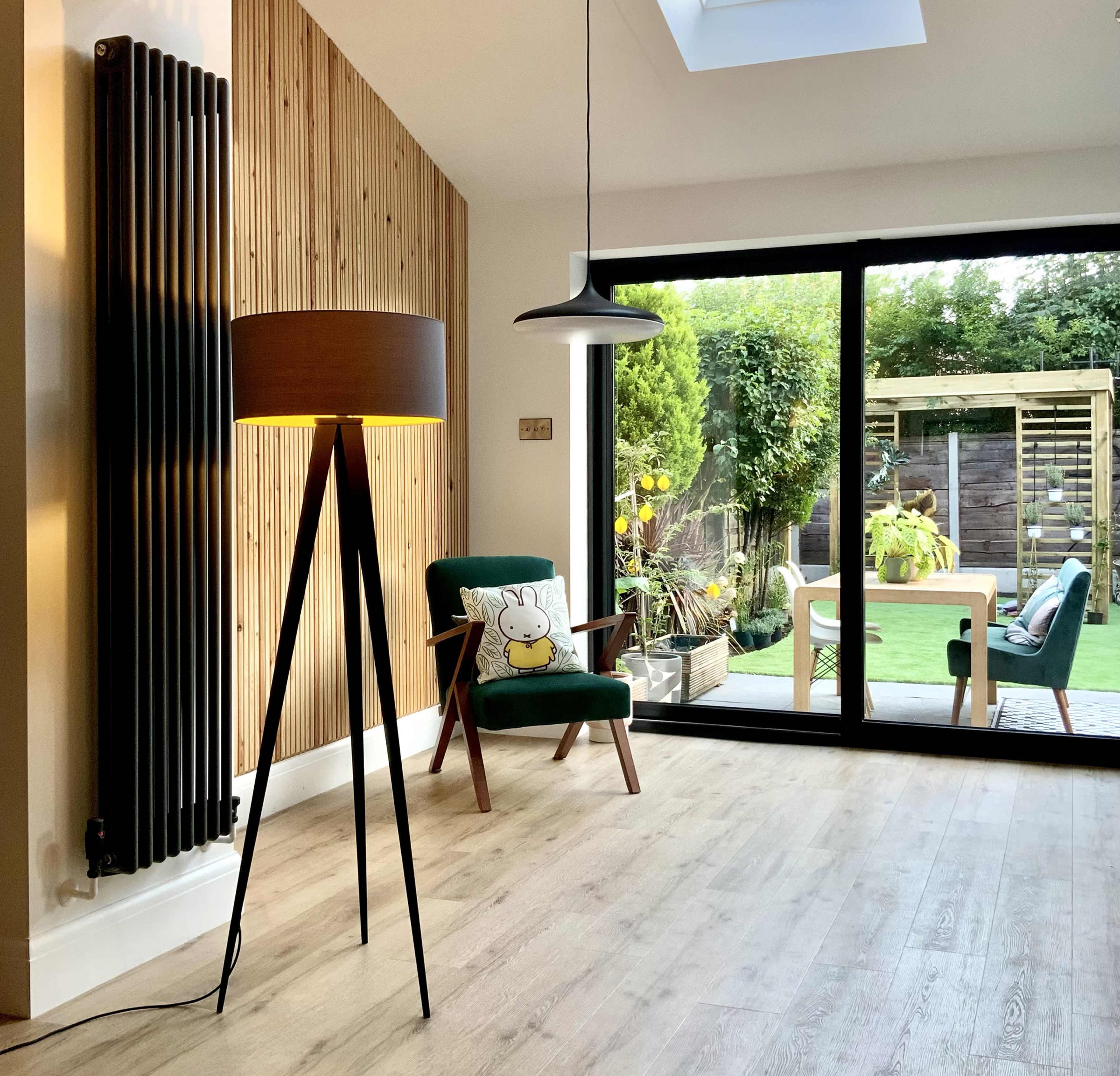 A modern living room features a tall, legged lamp and a green armchair with a decorative pillow, overlooking a garden through large glass doors.