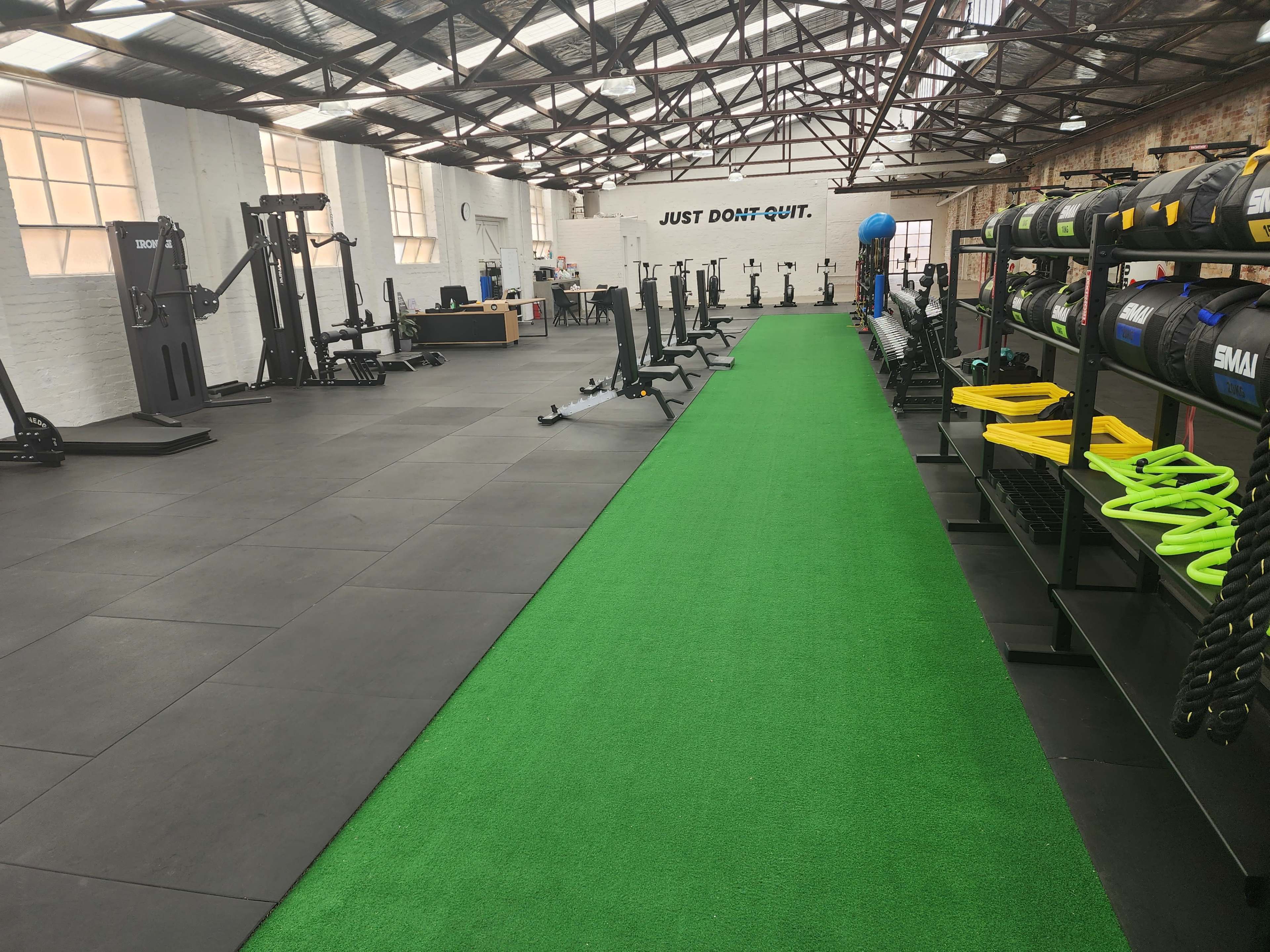The image shows a spacious gym with a green artificial turf area, various fitness equipment, and a motivational sign on the wall that reads "JUST DON'T QUIT."