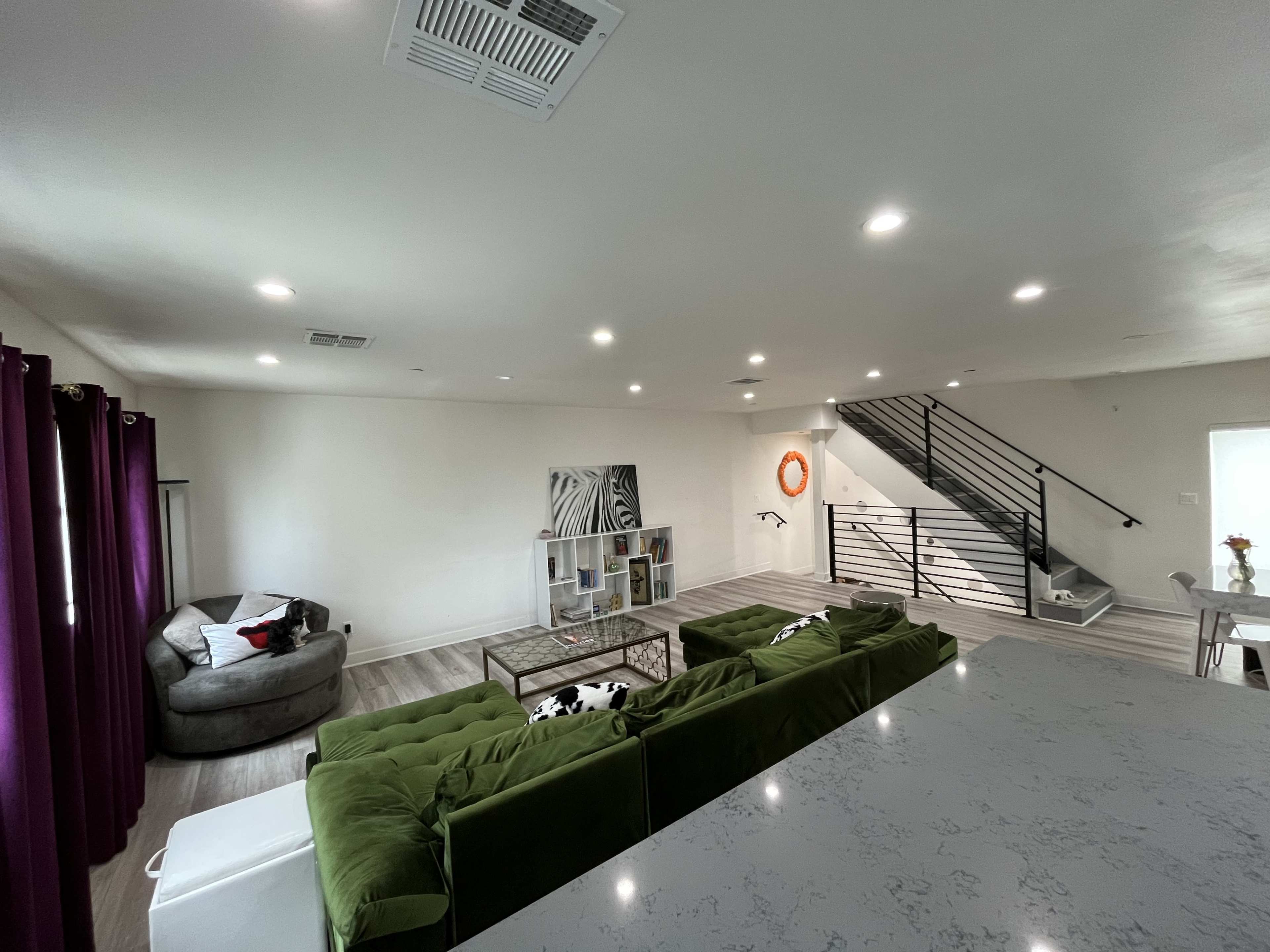 The image shows a modern living room with a green sectional sofa, a gray armchair, and a staircase with a metal railing in the background.