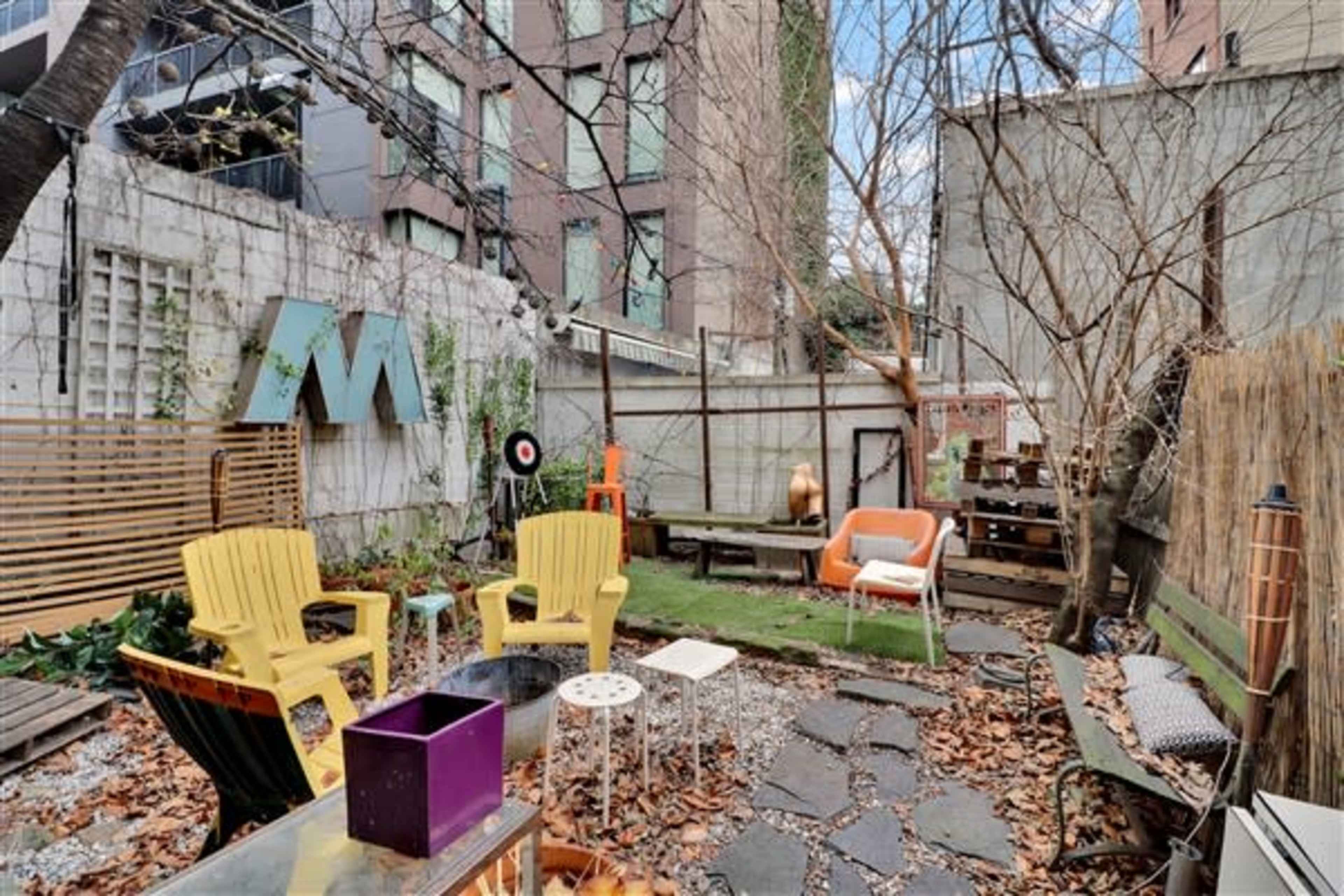 A small outdoor gathering area with yellow chairs, a wooden bench, and scattered leaves amidst urban buildings.