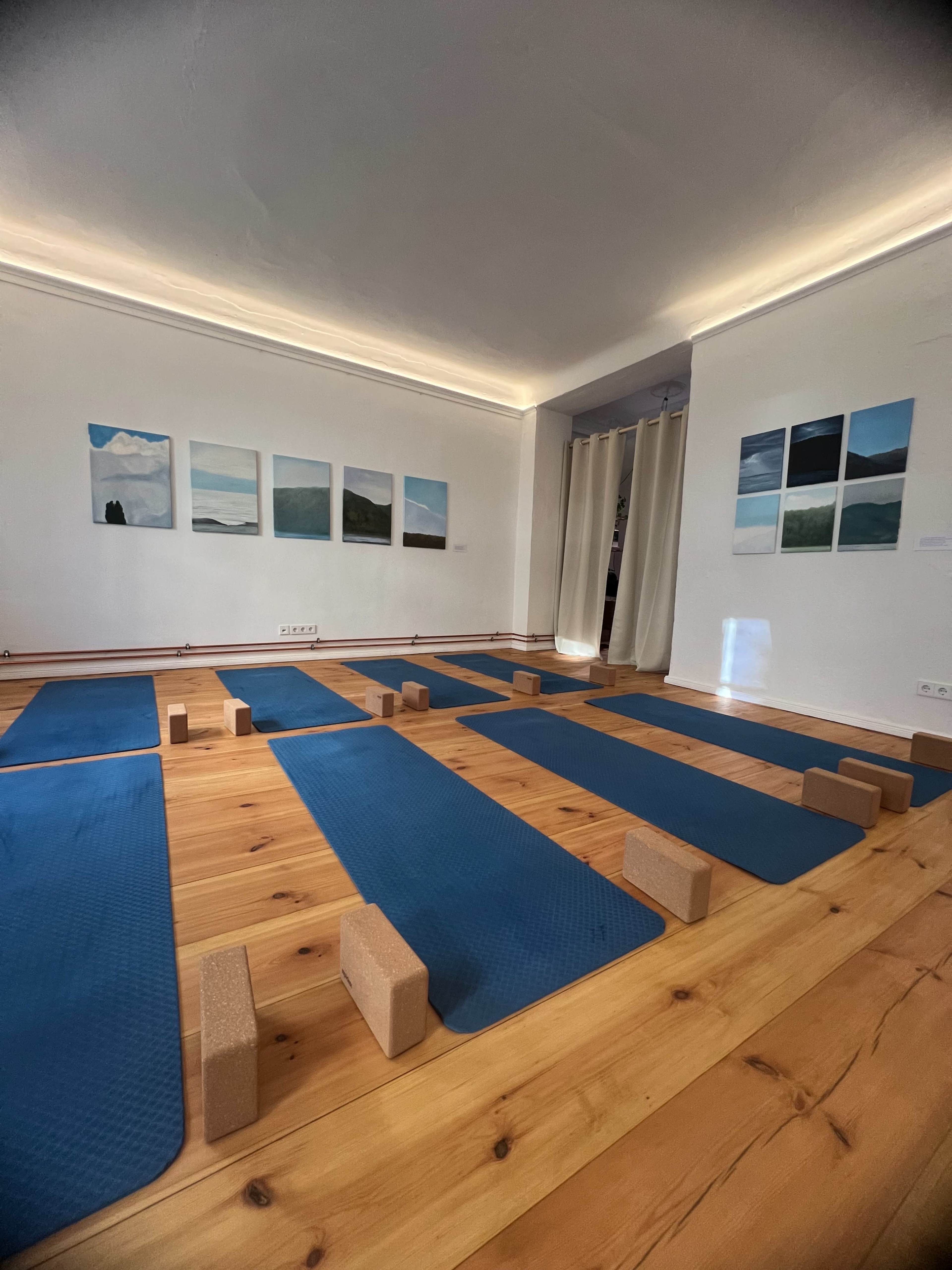 A yoga studio features several blue mats arranged on a wooden floor, with foam blocks placed on each mat and paintings on the wall.