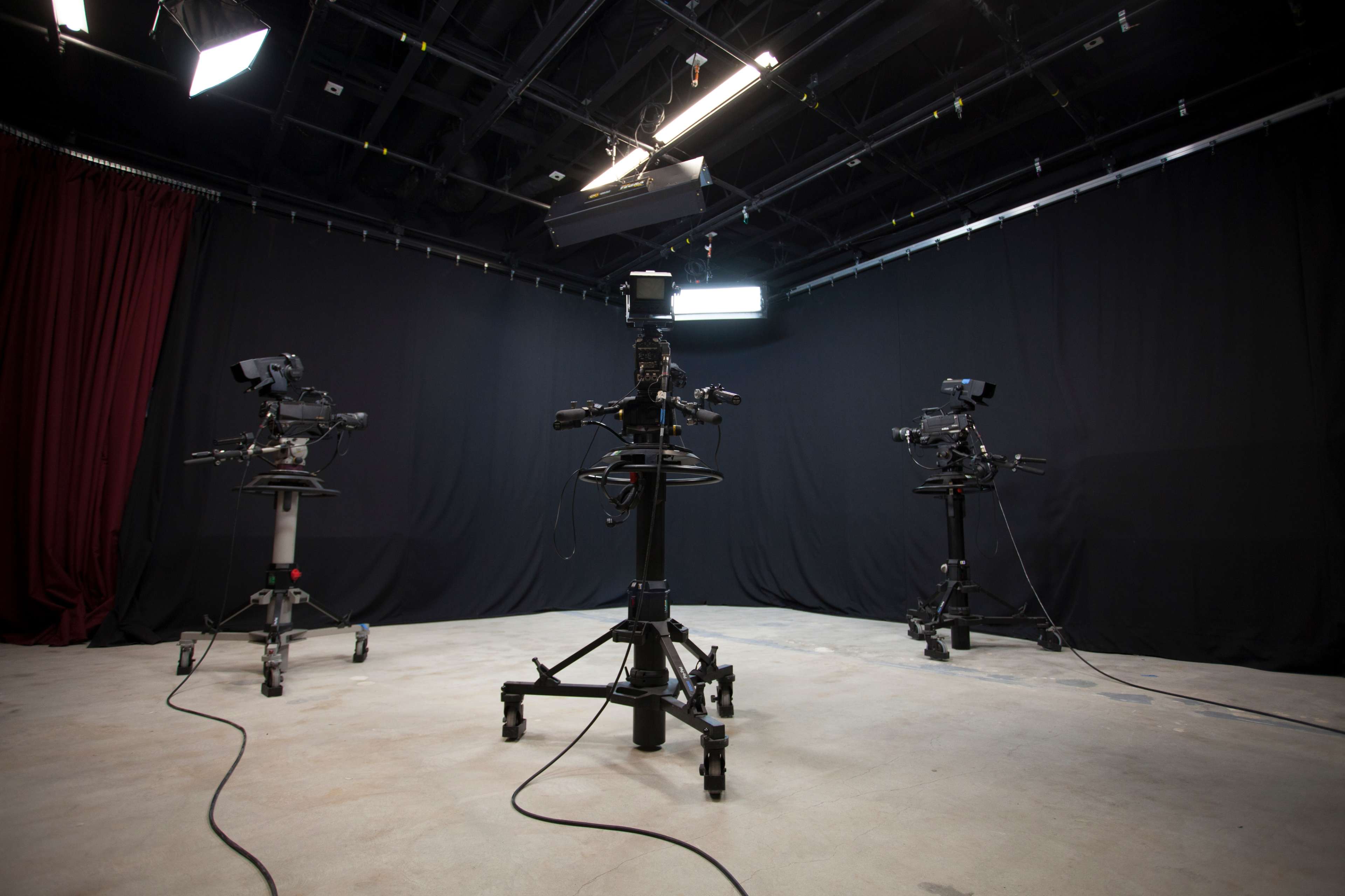 A television studio setup with three camera tripods facing a central point, surrounded by black curtains and overhead lighting.