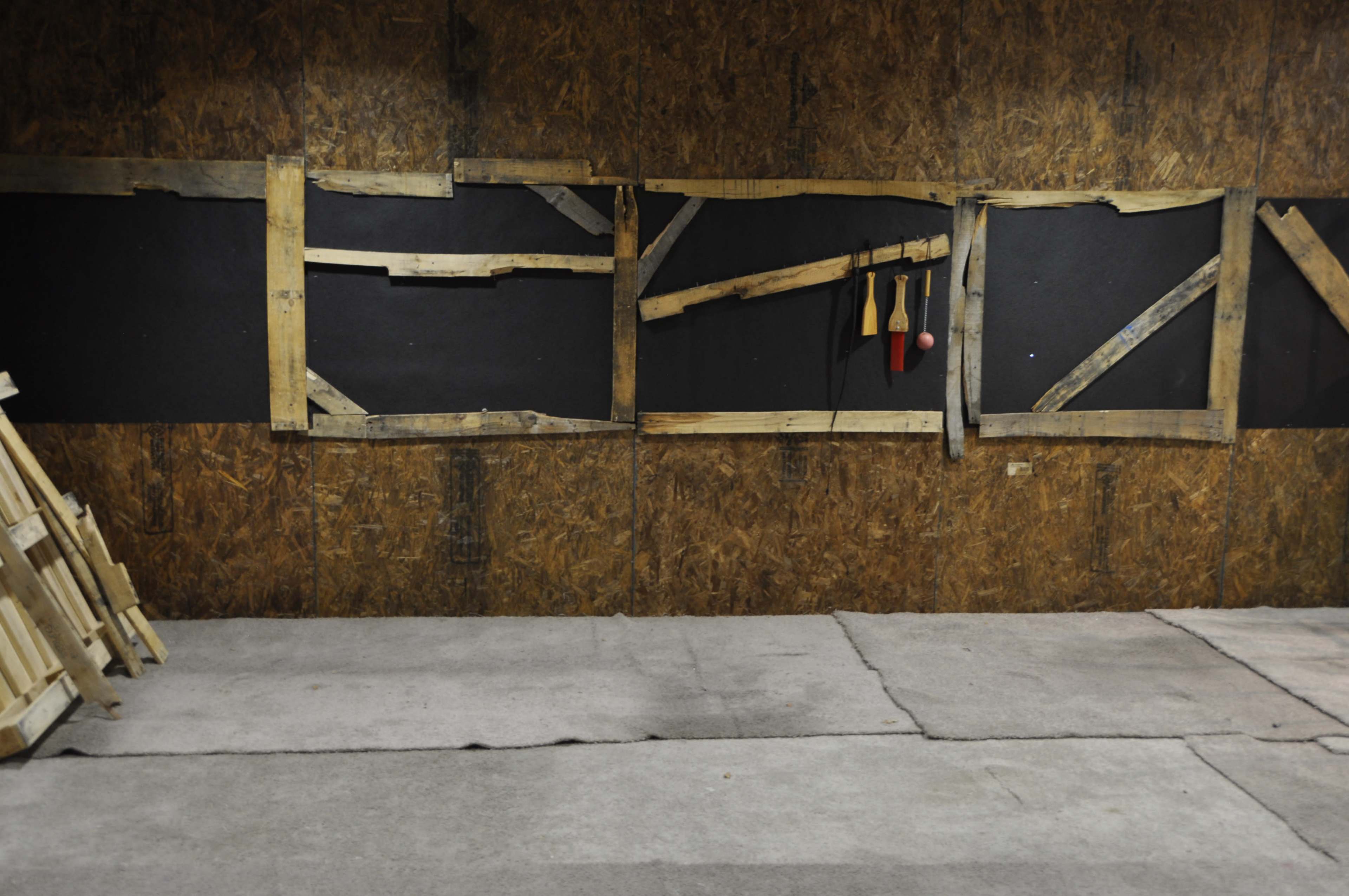 The image shows a wall made of textured wood panels with several wooden frames and tools hanging beside a concrete floor.