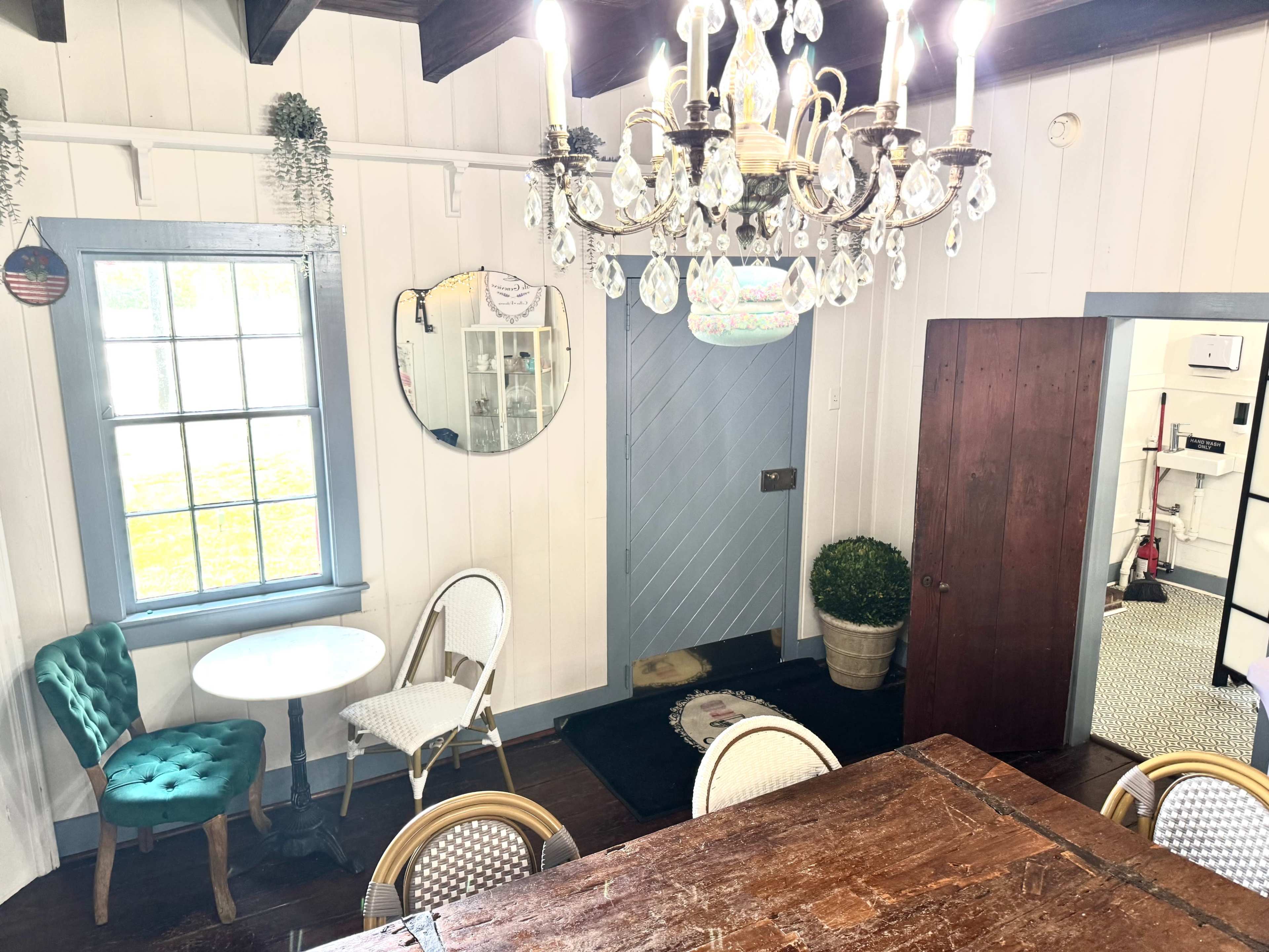 The image shows a cozy dining area featuring a wooden table, green velvet chairs, a chandelier, and a doorway leading to another room.