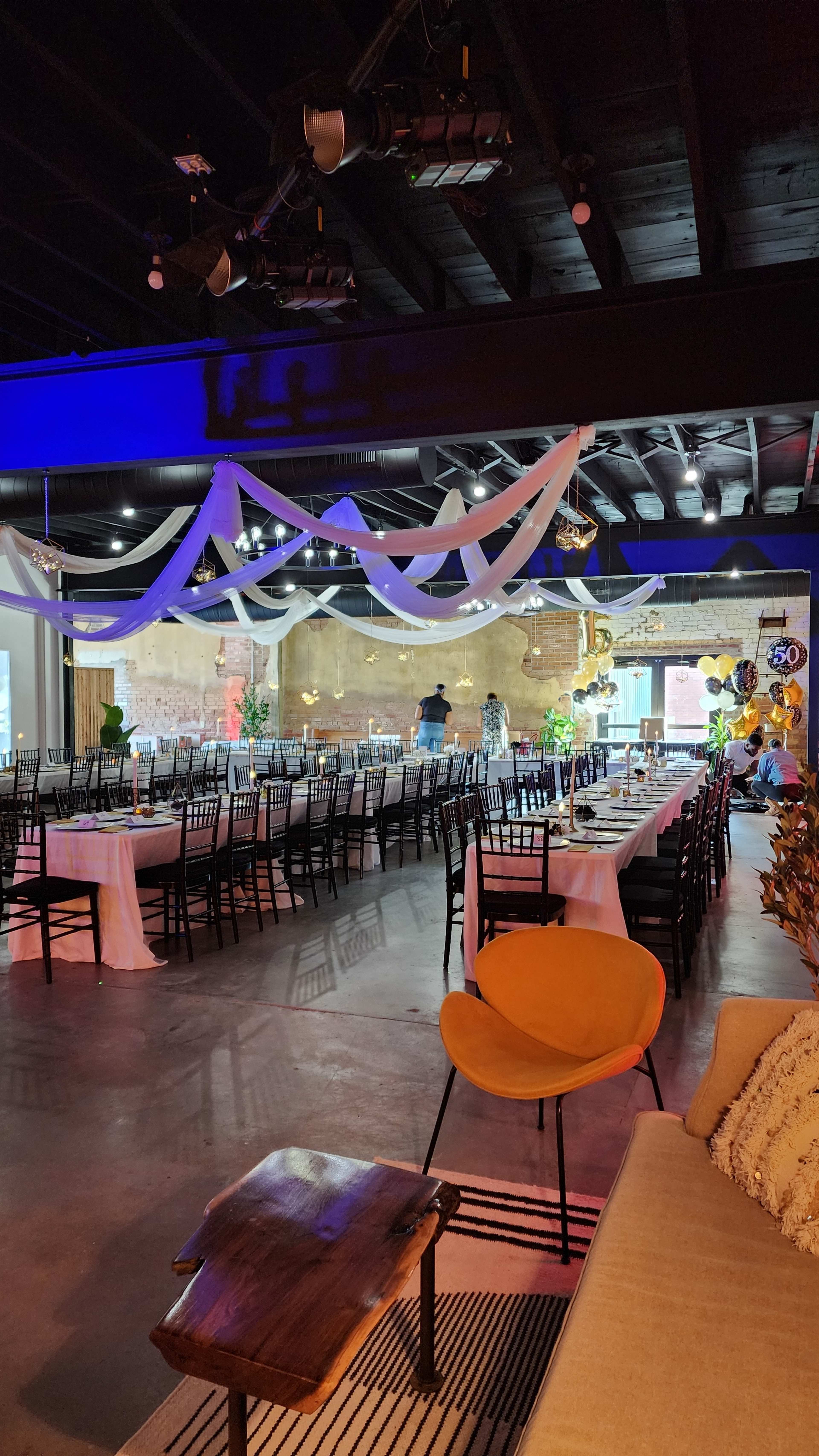 A spacious event venue features rows of neatly arranged tables and chairs under draped decorations and ambient lighting.