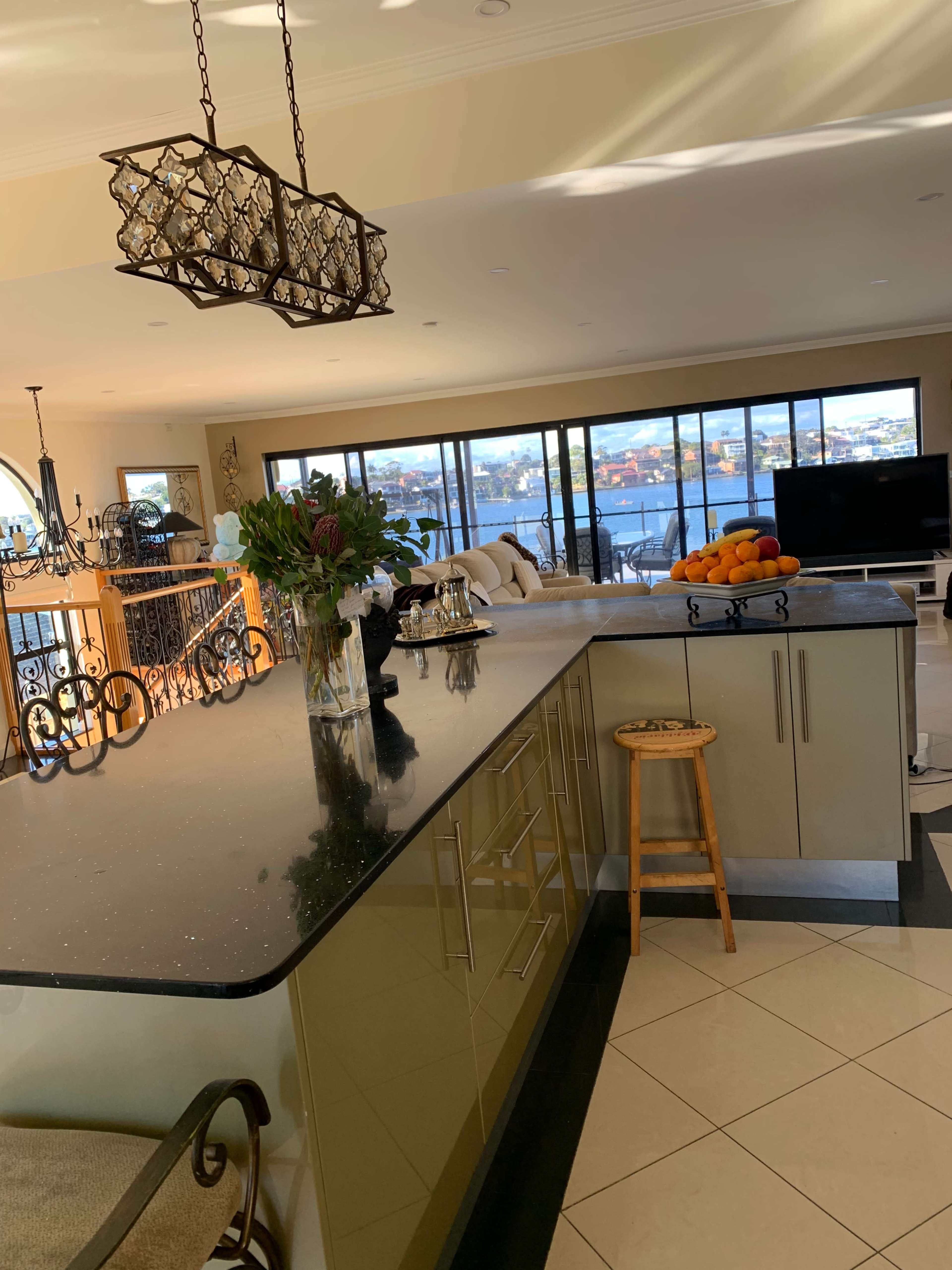 A modern kitchen with a dark countertop, a wooden stool, and large windows overlooking a waterfront view.