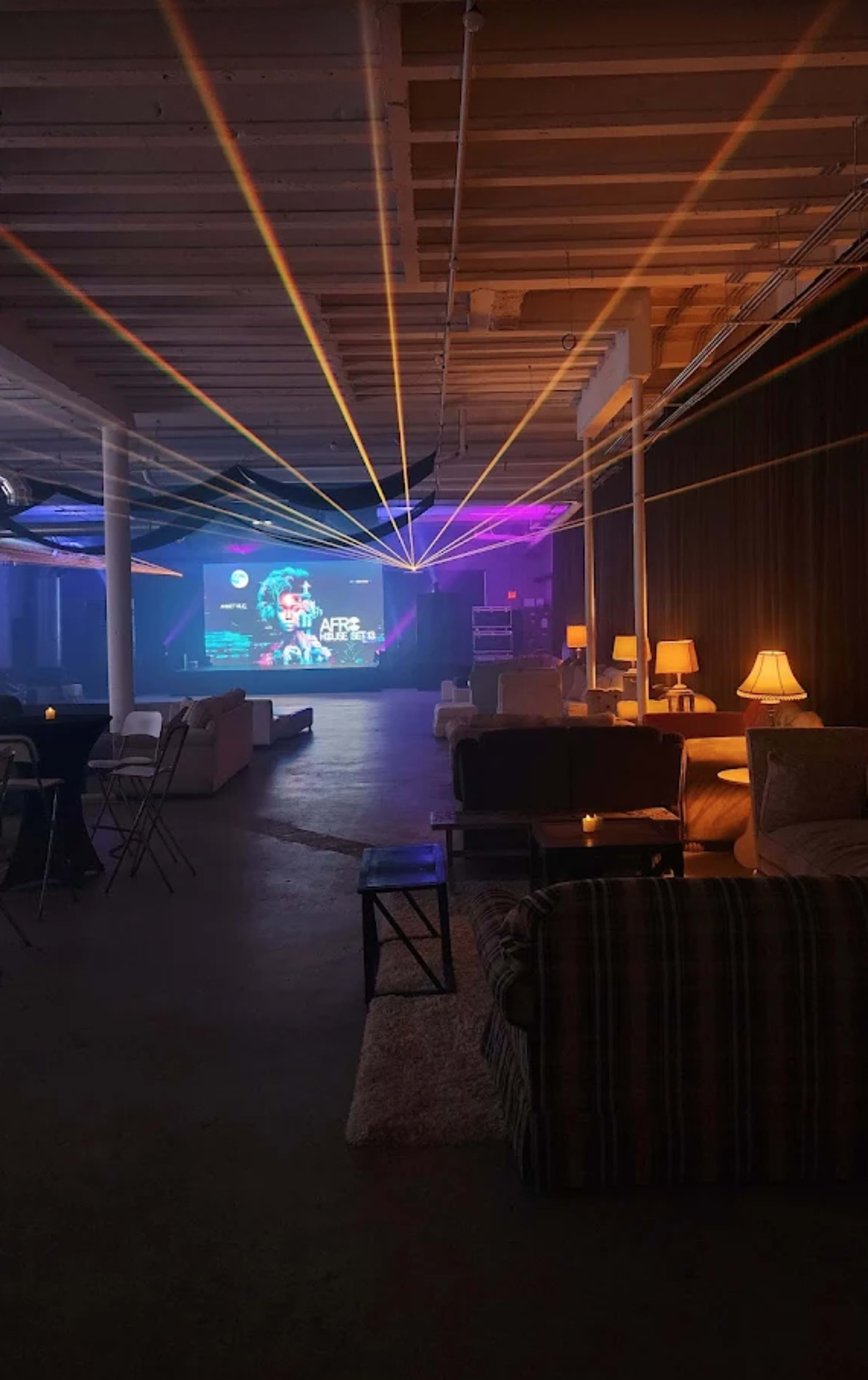 The image shows a dimly lit lounge area with comfortable seating, small tables, and colorful laser lights projecting onto a screen in the background.