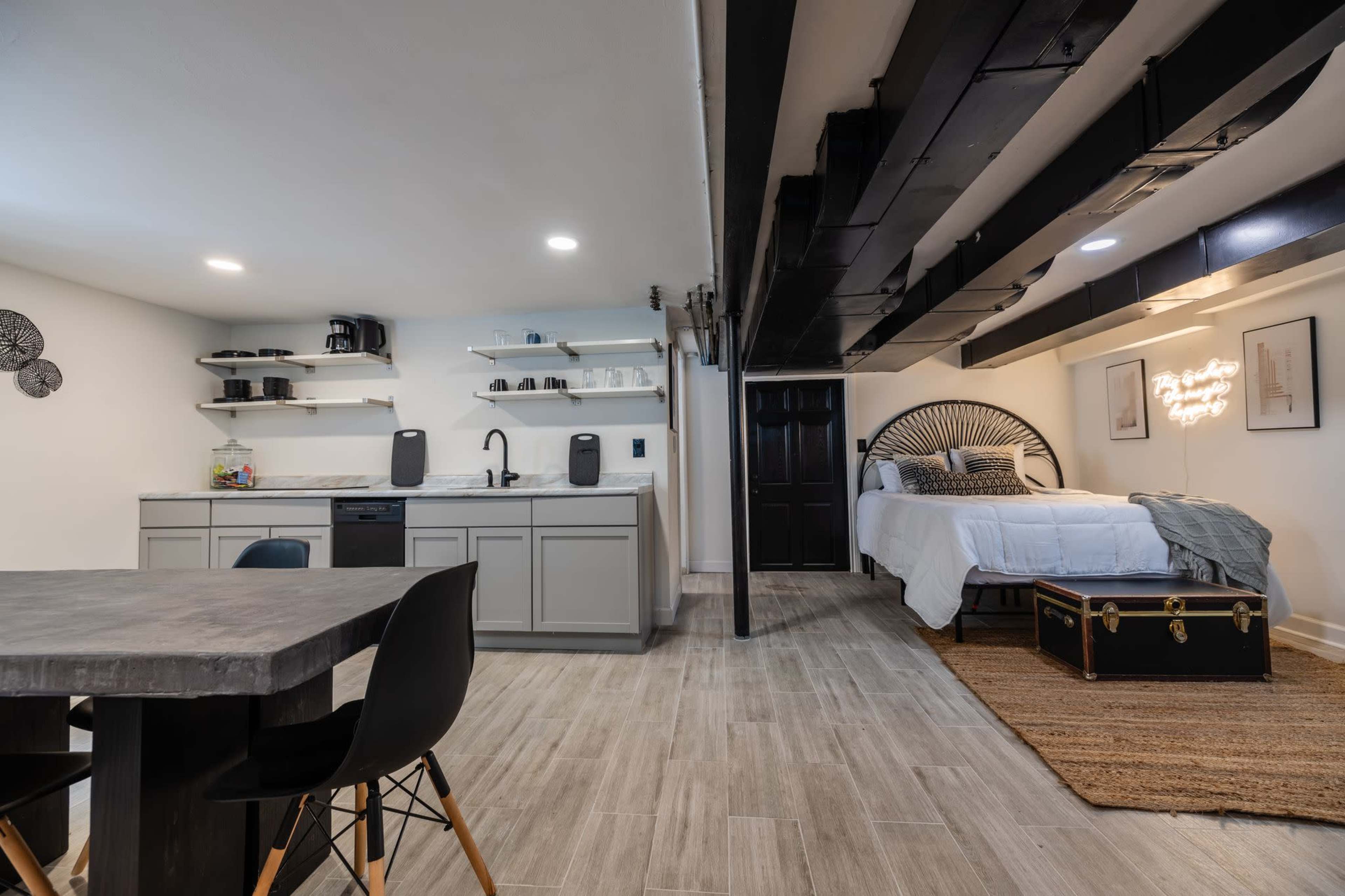 A modern basement studio features a kitchen area with open shelves and a dining table, adjacent to a sleeping area with a bed and decorative trunk.