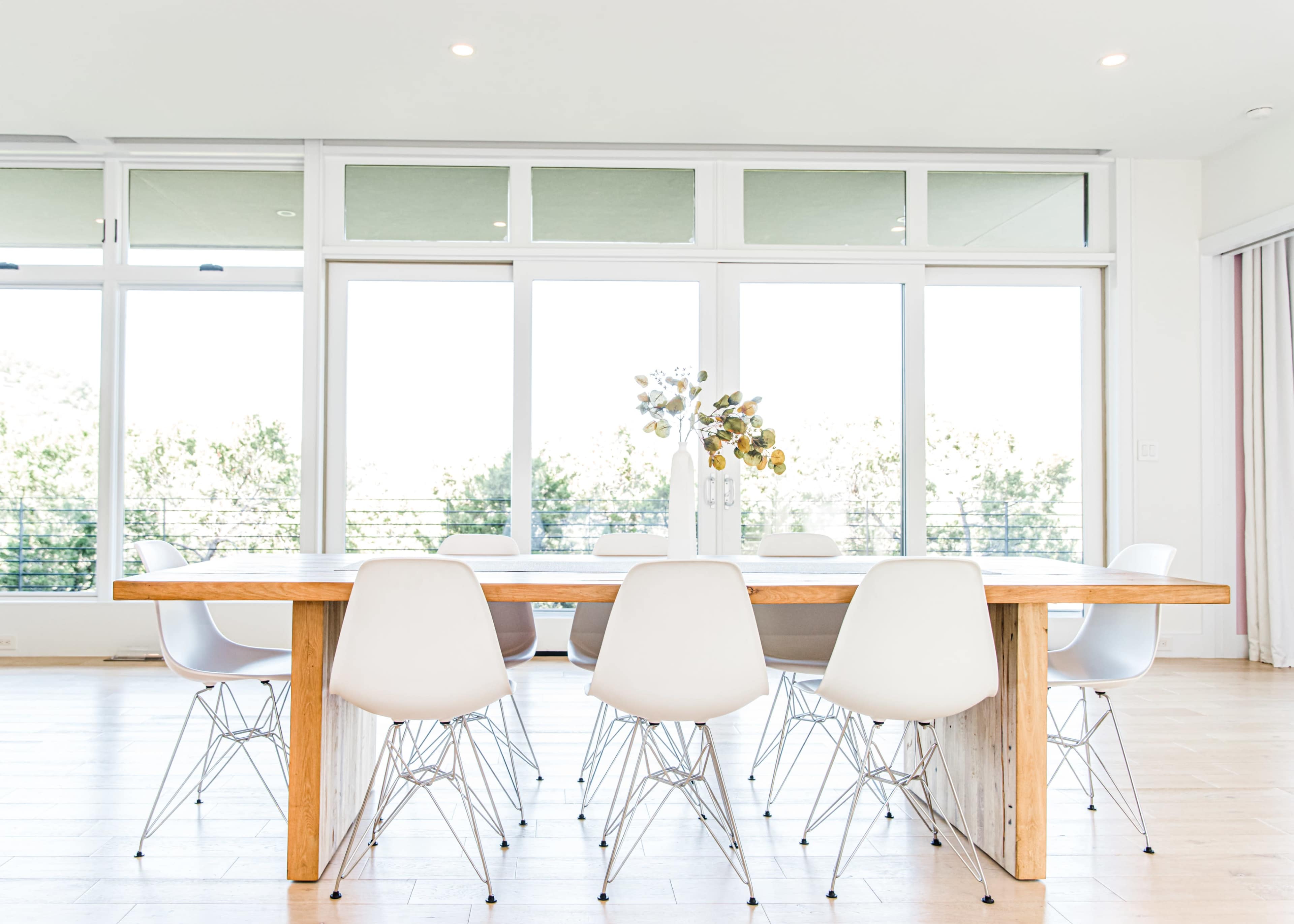 A large wooden dining table with white chairs is set in a bright room, featuring floor-to-ceiling windows and a view of greenery outside.