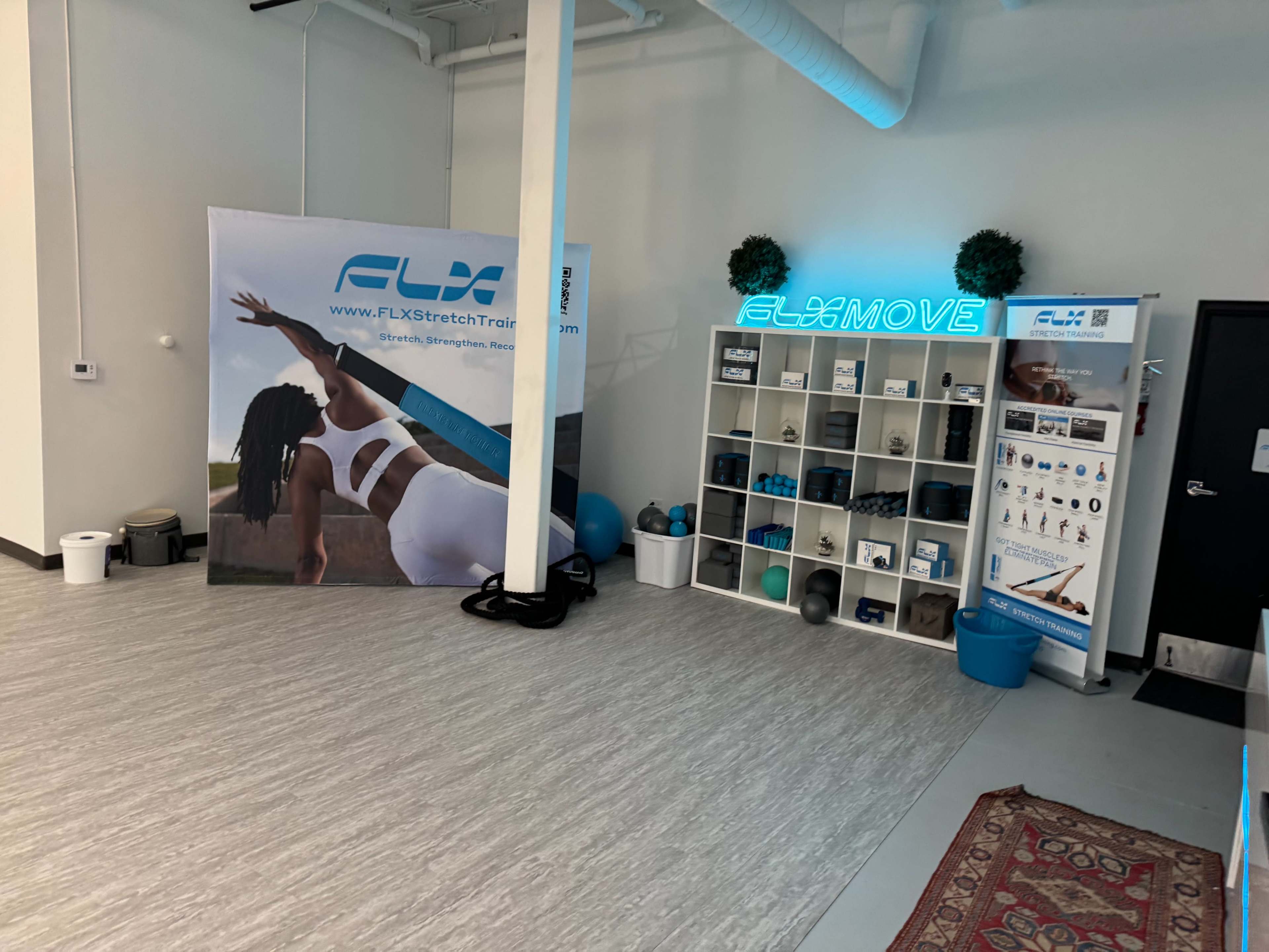 The image shows a fitness studio interior featuring a large banner for FLX Stretch Training and a shelving unit displaying various fitness equipment and informational materials.