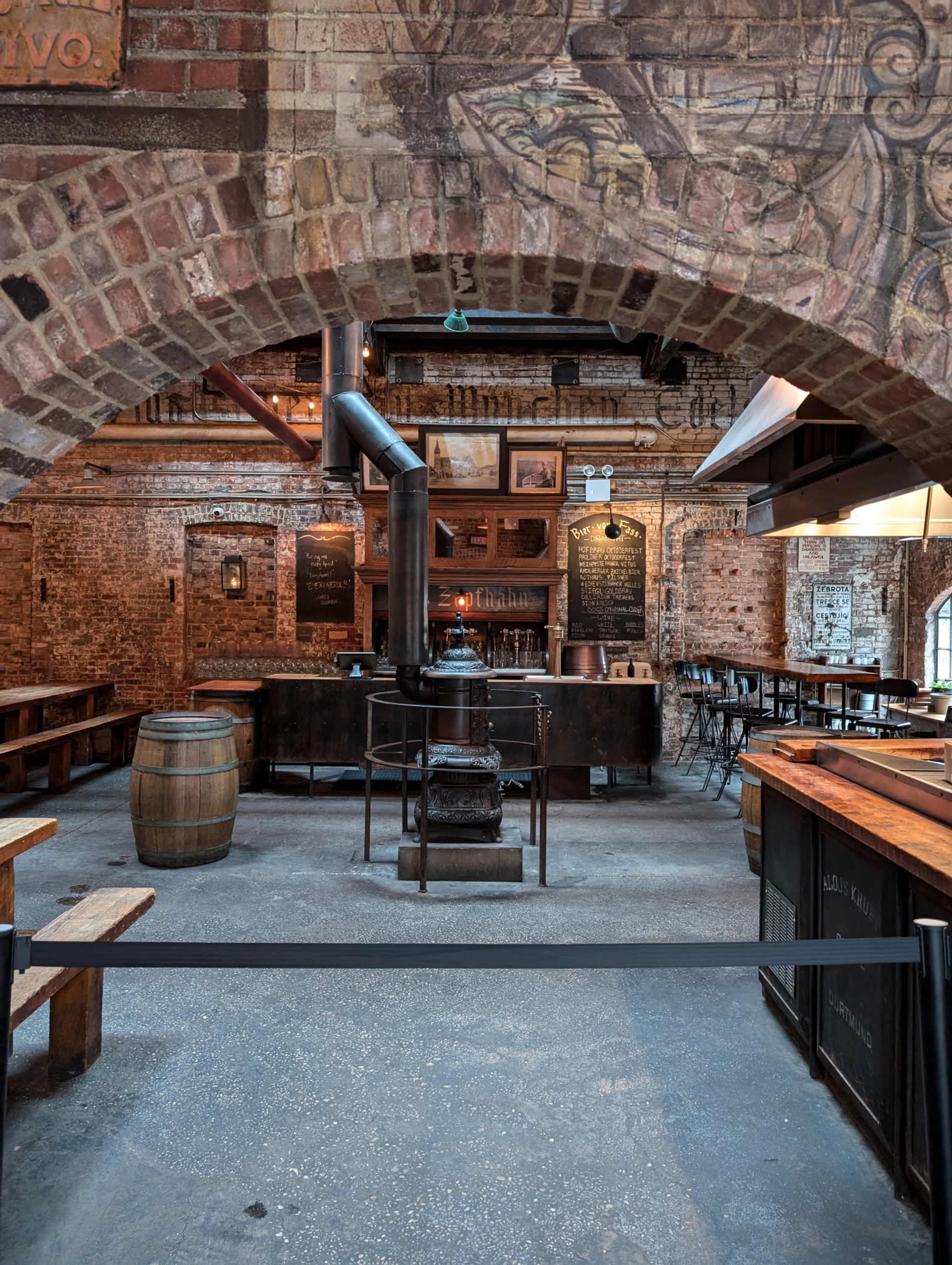 The image shows a rustic interior of a brick-walled brewery with wooden benches, barrels, and an industrial-style stove at the center.