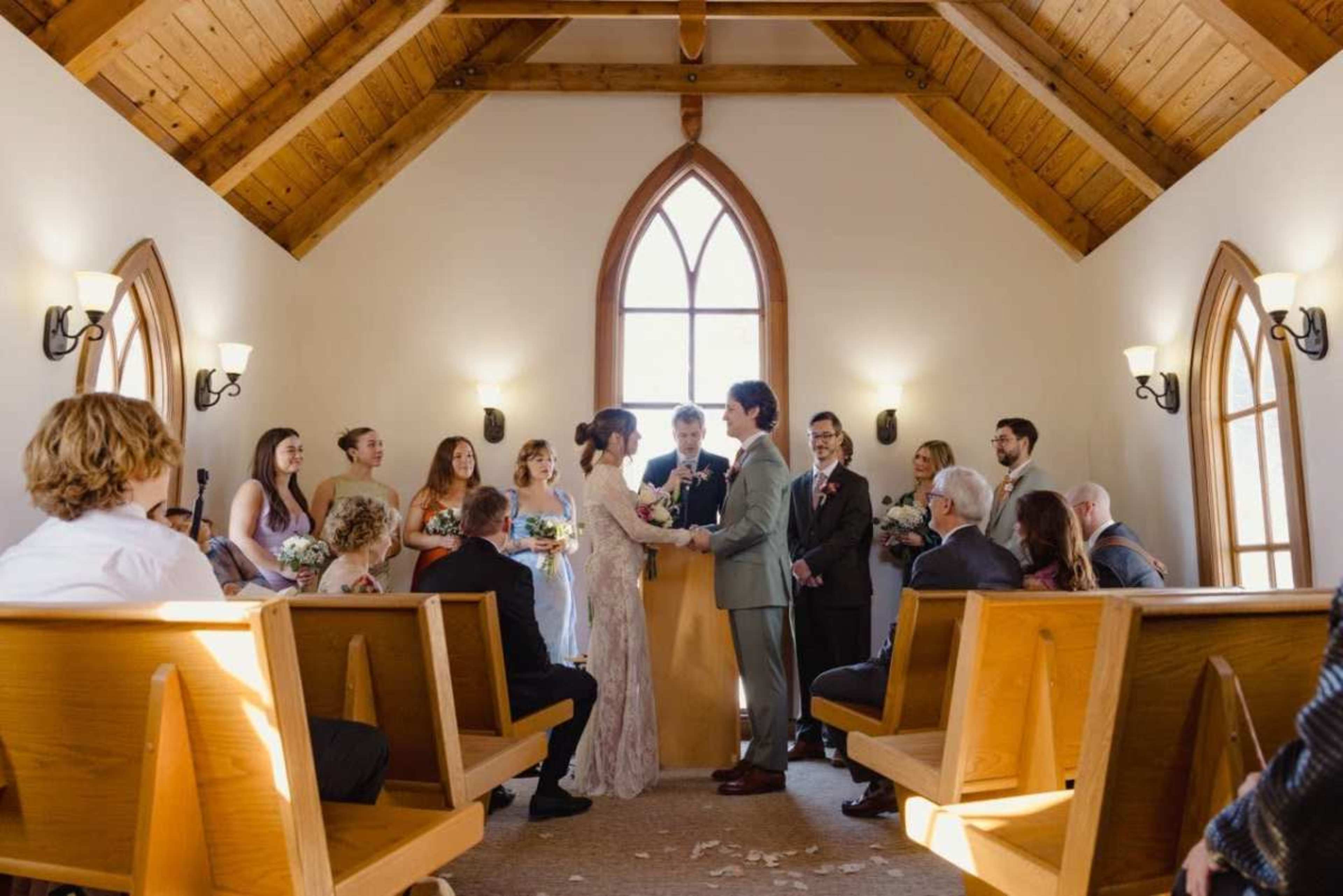 A couple stands at the front of a chapel, exchanging vows while surrounded by their wedding party and guests seated on wooden benches.